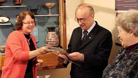 A woman in an orange jacket smiling while receiving an award or certificate from a man in a black suit, with a gray-haired woman nearby observing, inside a room with decorative plates and vases.