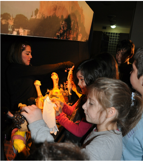 Children at a puppet show, watching puppets and a performer.