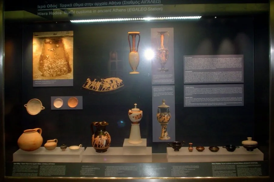 Display case featuring ancient Greek pottery and artifacts, with informational text panels in the background.