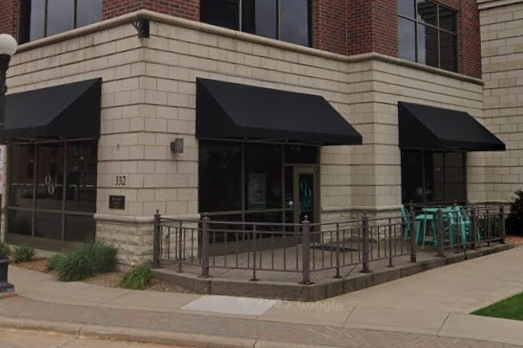 Corner of a building with black awnings over windows and door, with outdoor seating including teal chairs, railing around the patio, and sidewalk in front.