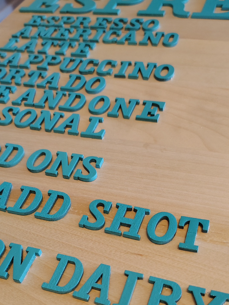 Blue plastic letter tiles arranged on a wooden surface, forming words related to coffee and beverages, including 'ESPRESSO', 'AMERICANO', and 'LATTE'.