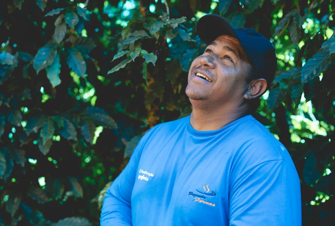 Man in a blue shirt and black cap smiling while standing outdoors among lush green leaves.