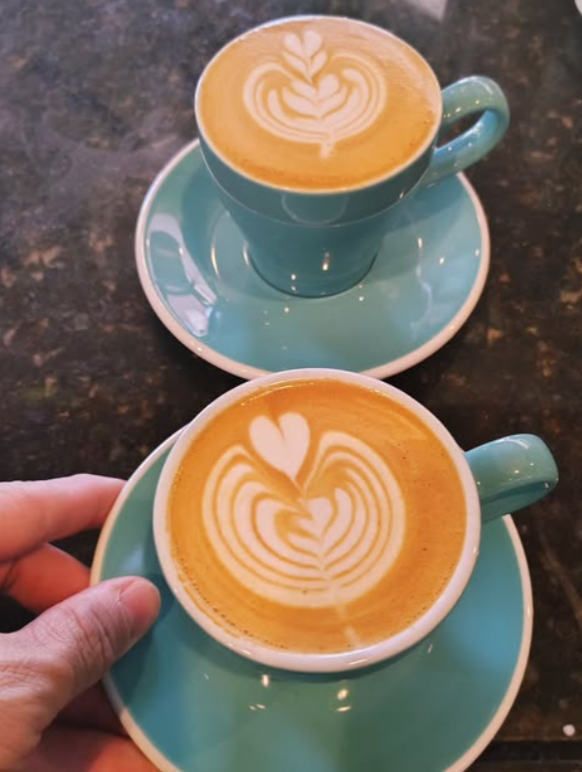 Two blue cups of coffee with latte art depicting hearts and swirls, sitting on matching blue saucers on a dark table.