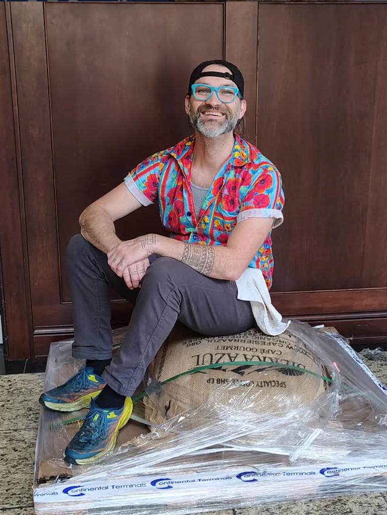 A man with glasses and a beard wearing a colorful floral shirt, gray pants, and sneakers, sitting on a burlap sack that is inside a plastic wrap, in front of a wooden wall, smiling at the camera.