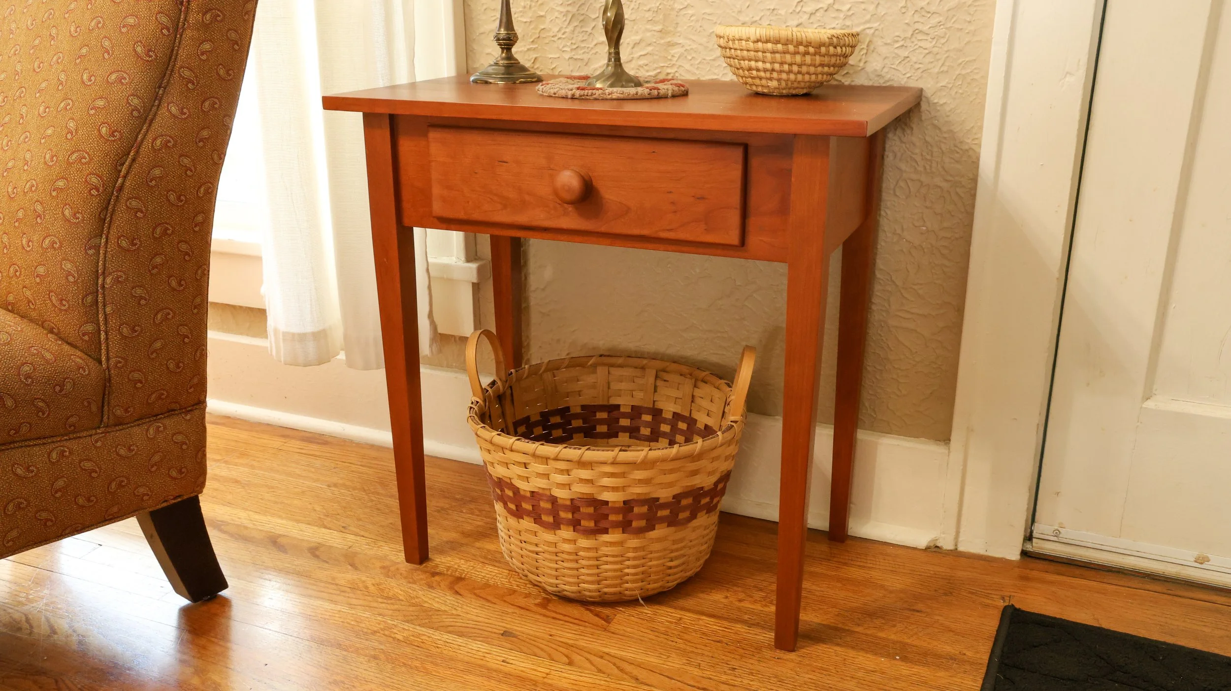 Wooden side table with drawer, wicker basket underneath, wicker bowl on top, hardwood floor, patterned upholstered chair nearby, white curtain.