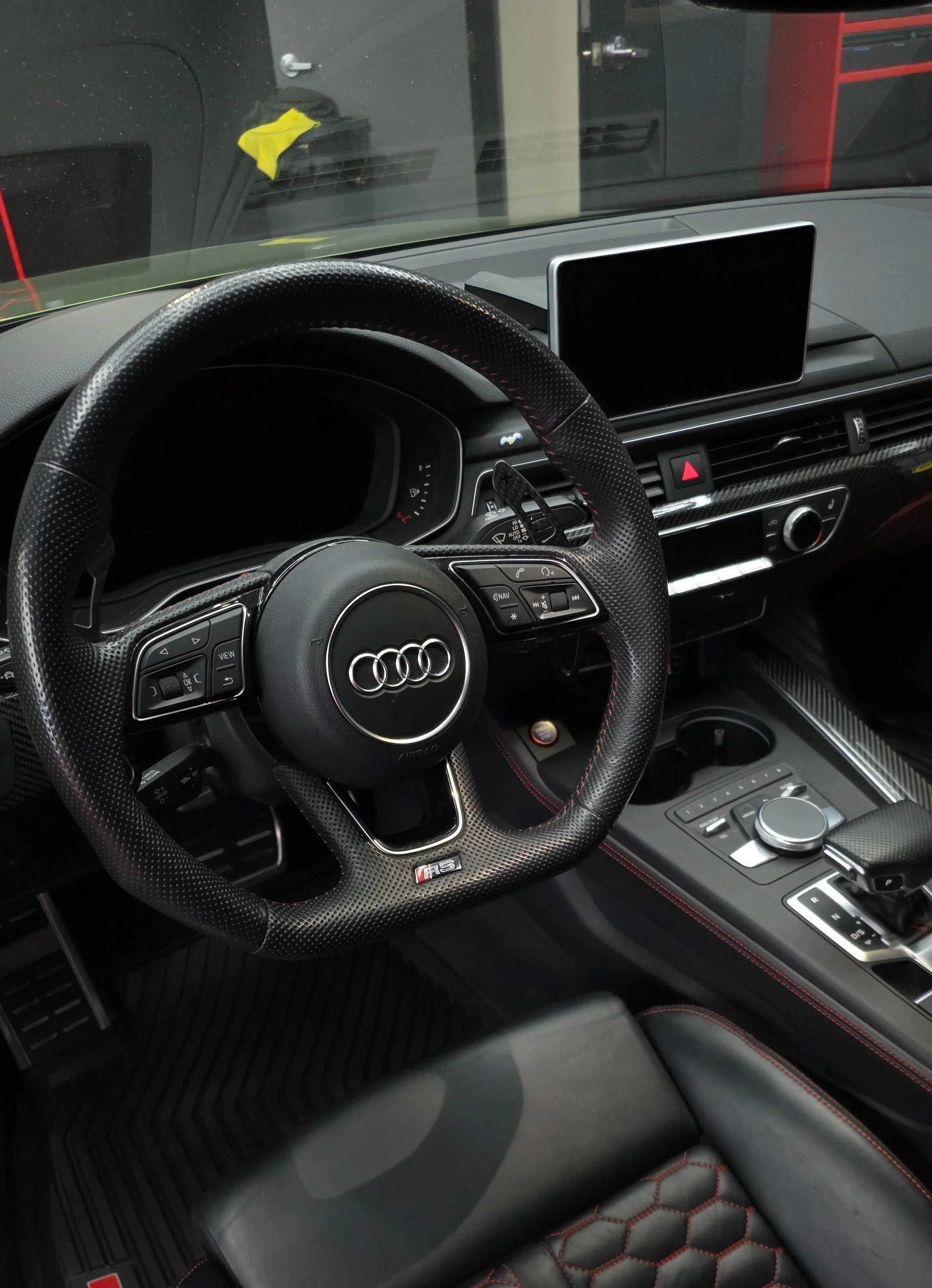 Interior of an Audi sports car showing a black perforated steering wheel with Audi logo, digital dashboard, display screen, control buttons, and a gear shifter with a rotary dial, all with black and red accents.