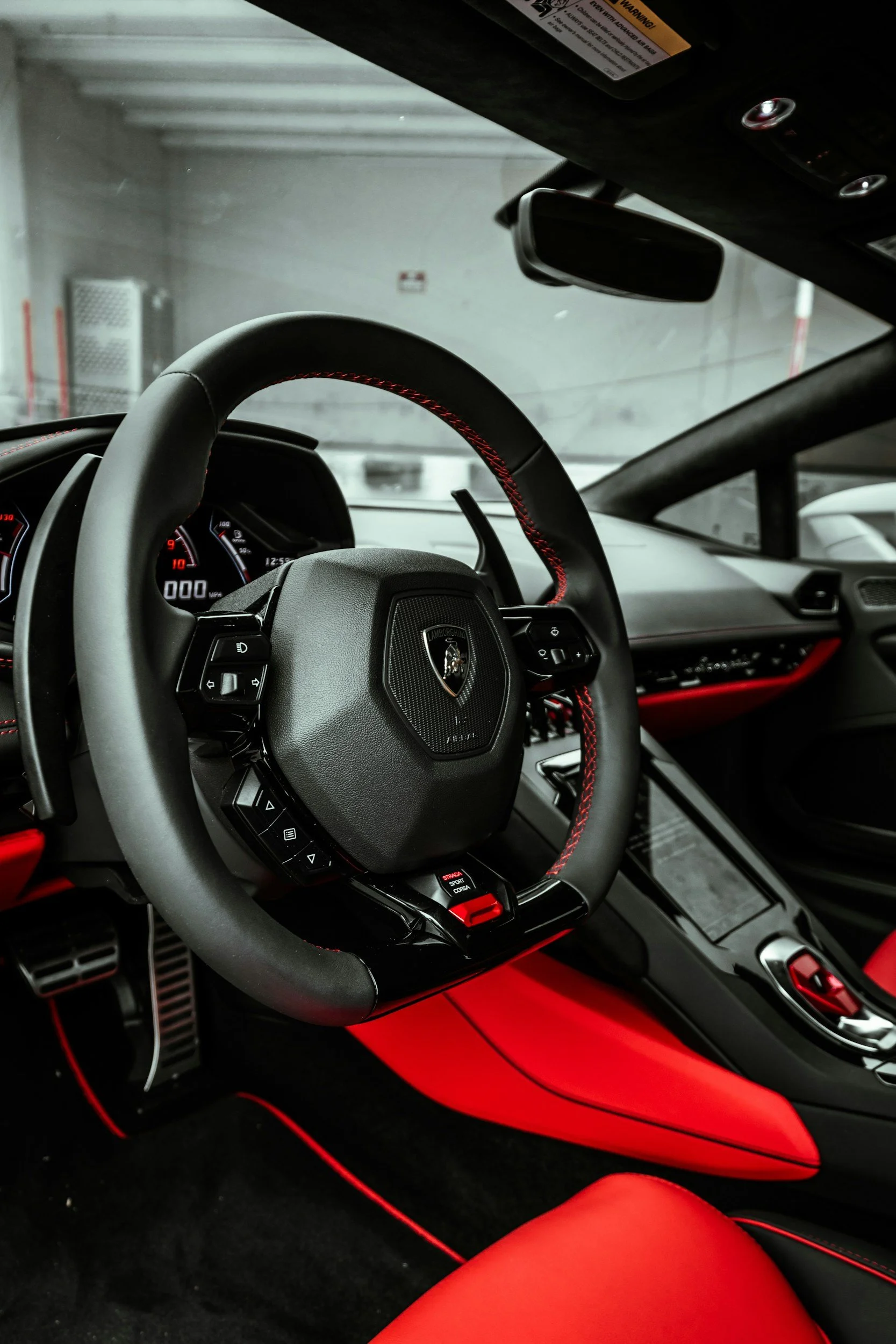 Interior of a Lamborghini car, showing the steering wheel with the Lamborghini logo, digital dashboard, and red and black interior accents.