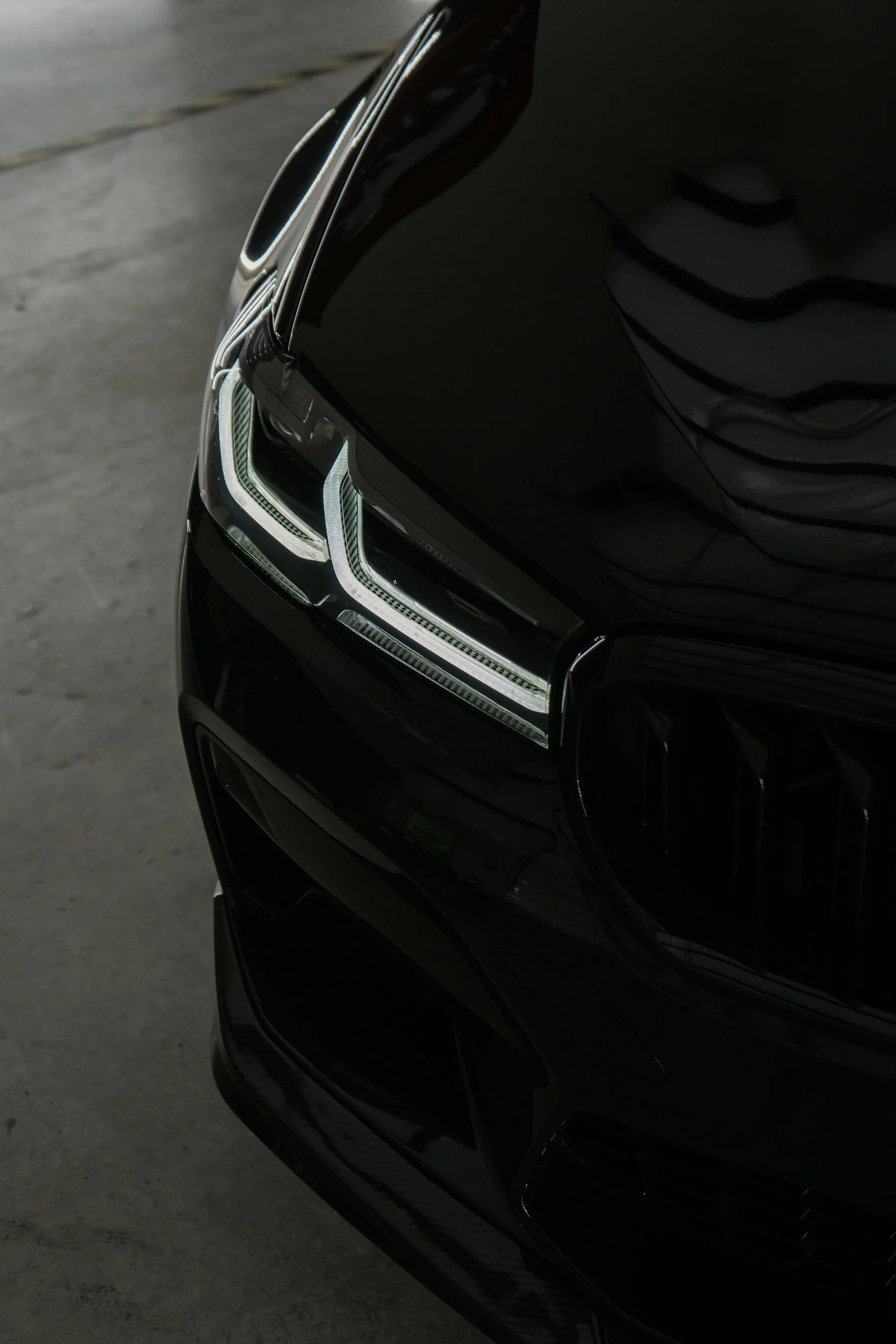 Close-up of a black luxury car's front, showing the right headlight with LED accents and a part of the hood, reflected on a smooth concrete surface.