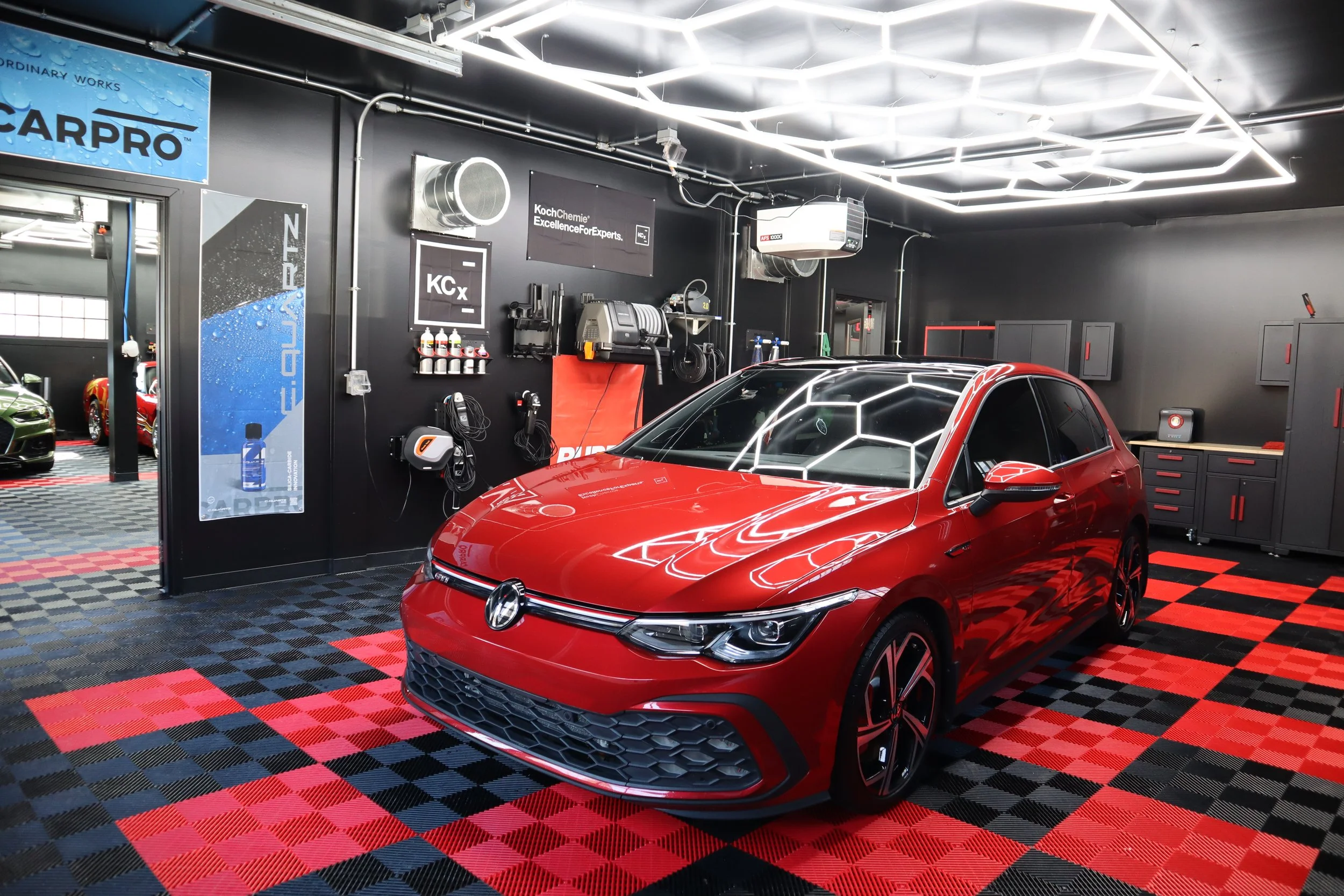 Red Volkswagen car inside a modern garage or detailing shop with black and red checkered flooring and black walls, various tools and equipment on the walls, and cars visible through an open door.