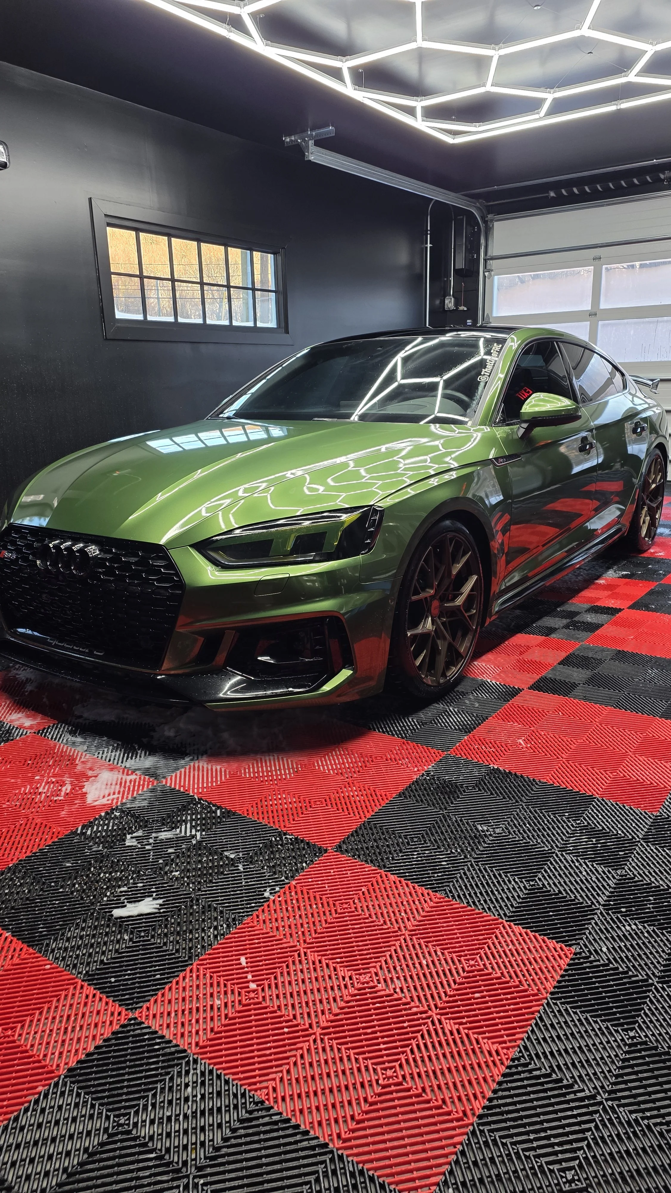 Green Audi sports car inside a garage with black and red checkered flooring and black walls, illuminated ceiling lights, and a window with sunlight outside.