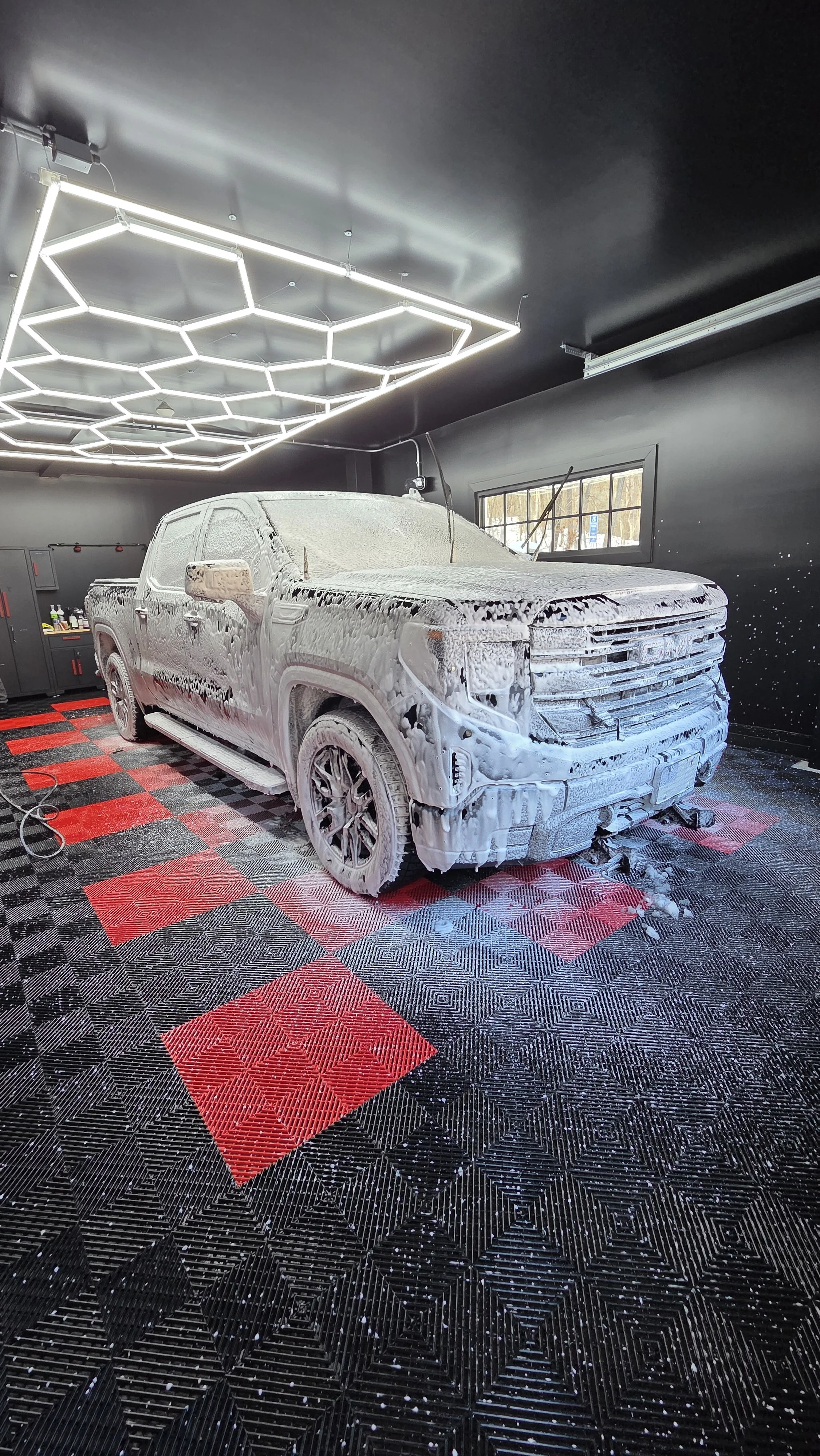 A white pickup truck covered in soap foam inside a car wash bay with a black and red checkered floor, and modern hexagonal LED ceiling lights.