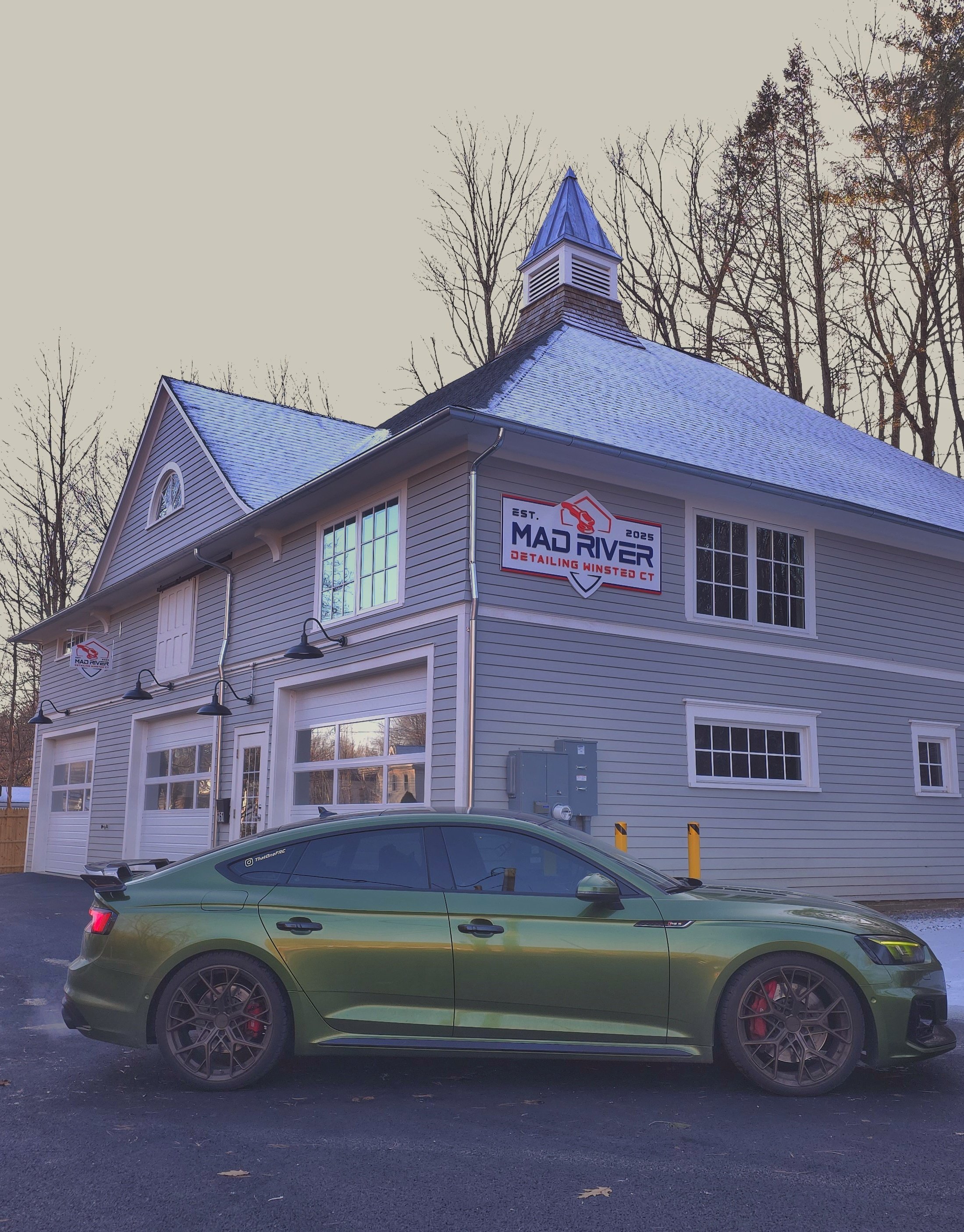 A green sports car parked in front of a light blue, two-story building with white trim and siding. The building has a small steeple and multiple signs for Mad River Detailing, Winsted, CT. Trees without leaves are visible in the background.