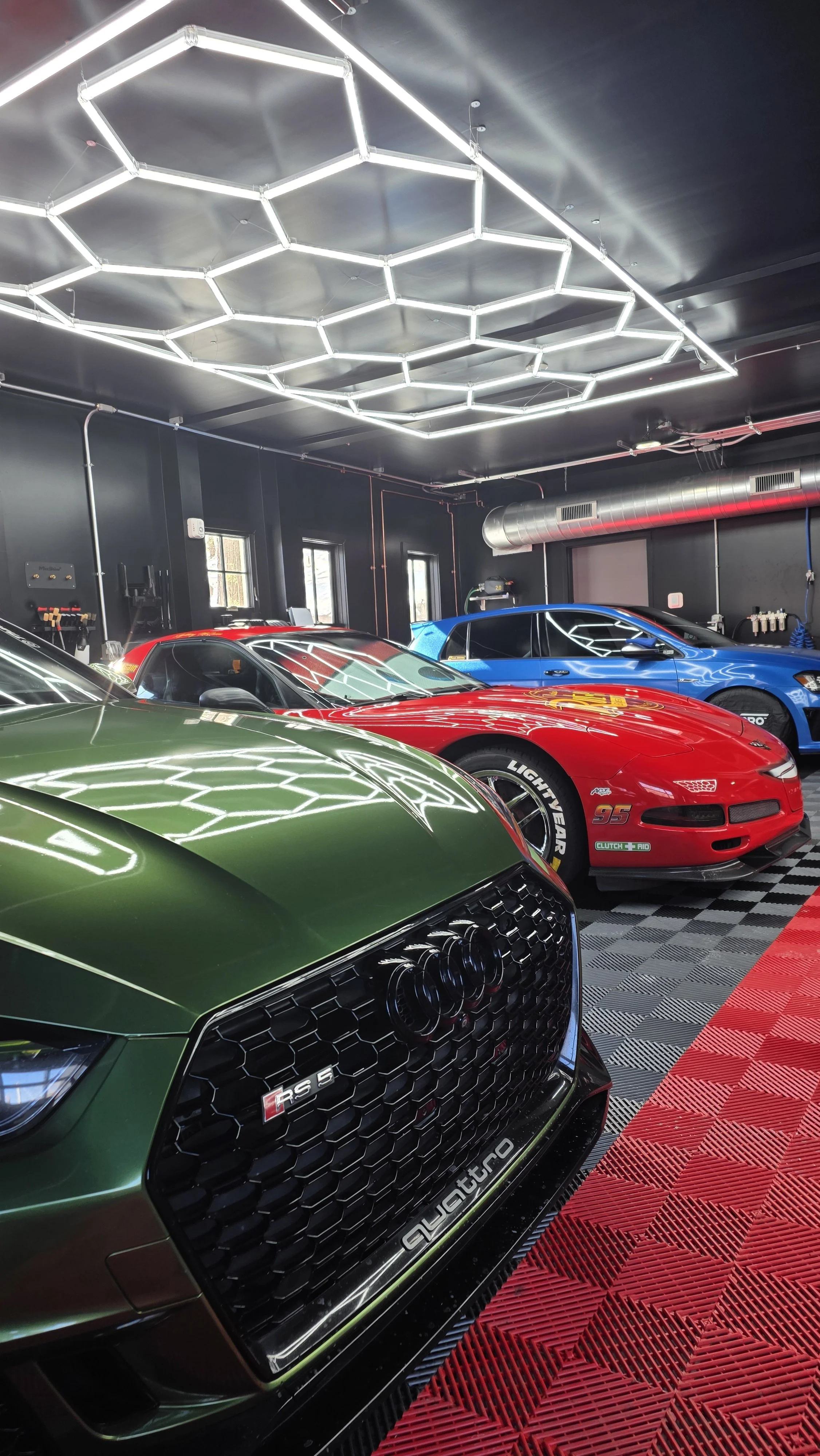 Interior of a garage with three cars: a green Audi RS 5, a red NASCAR style race car, and a blue sports car, illuminated by modern geometric LED ceiling lights.