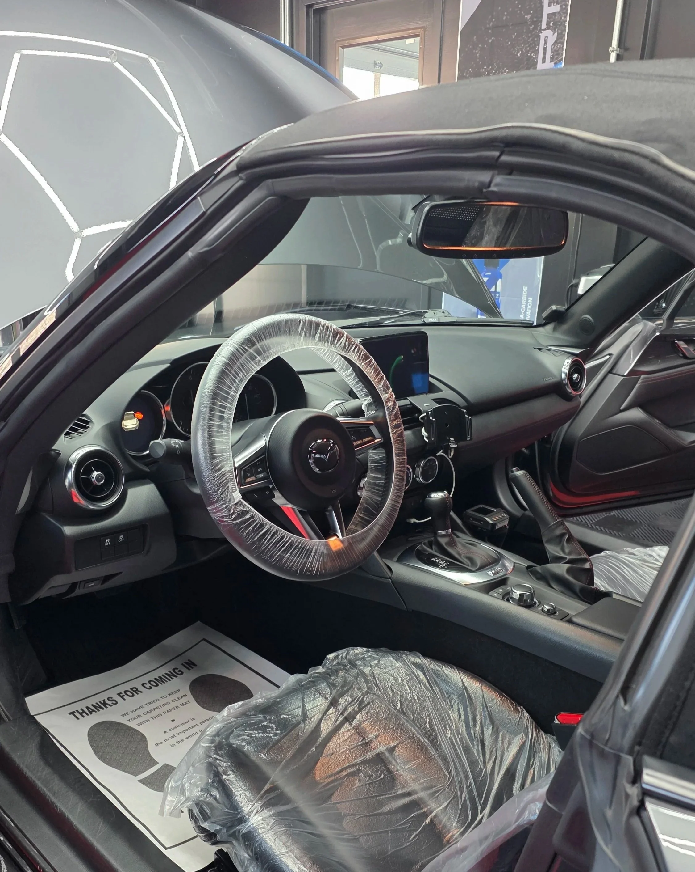 Interior of a new Mazda convertible car with plastic wrapping on the steering wheel and seat, and a paper floor mat that reads "Thanks for Coming In." The dashboard features a digital display screen, air vents, and controls, with the top down revealing the outside environment.