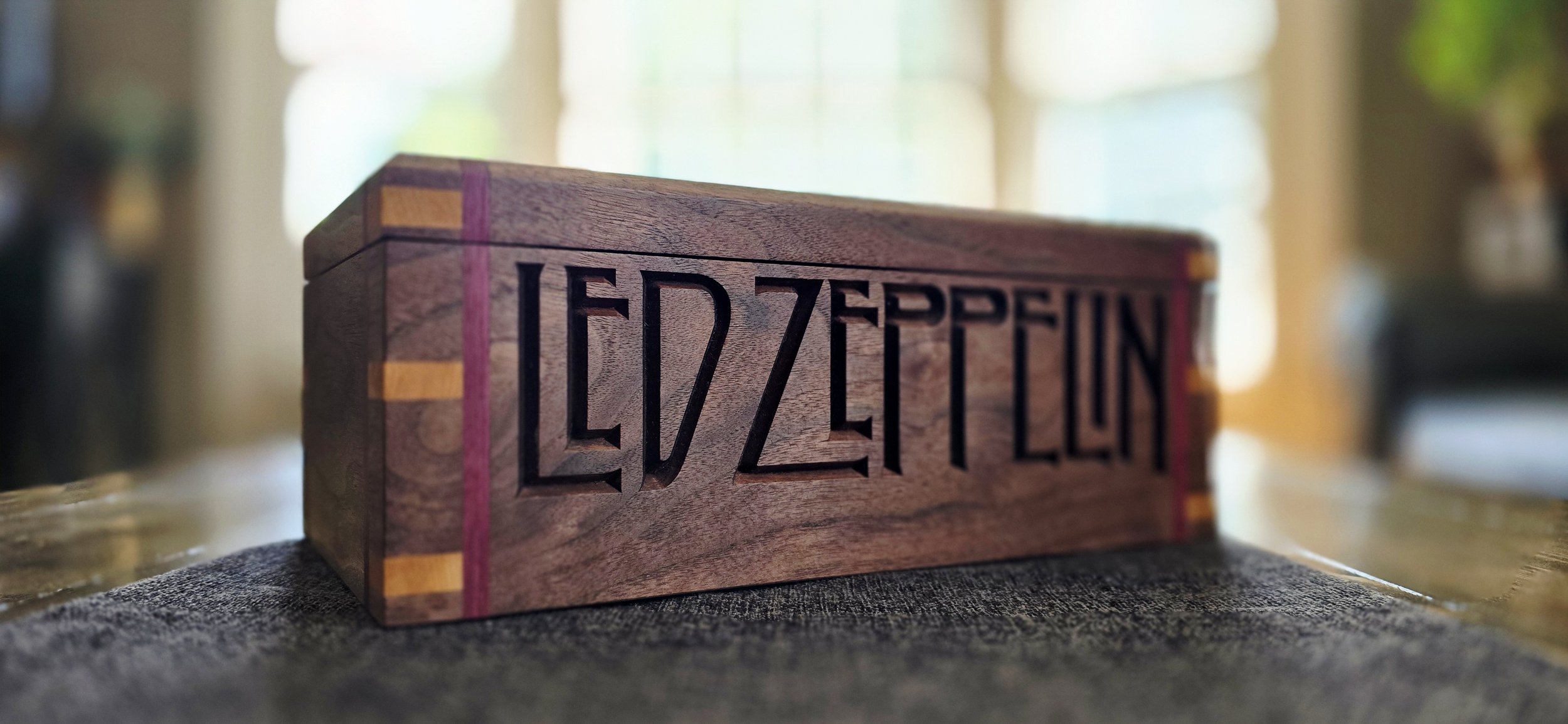 A wooden Lézard Lecle board with engraved text, placed on a dark fabric surface, with a blurred background of indoor space and windows.