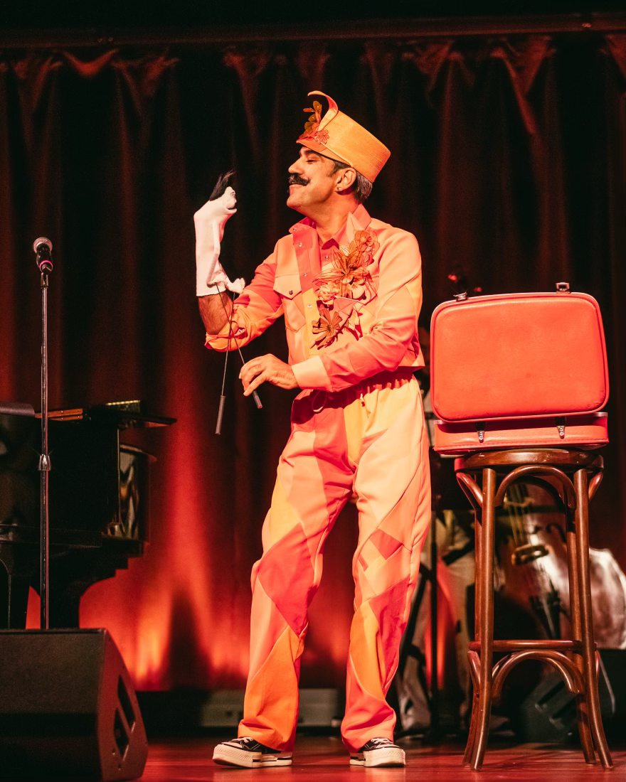 A man dressed in a colorful, floral-themed costume with an orange hat and pants, performing on stage with a puppet on his hand. The stage background has red curtains, and there is a stool with a red seat and a black piano in the background.