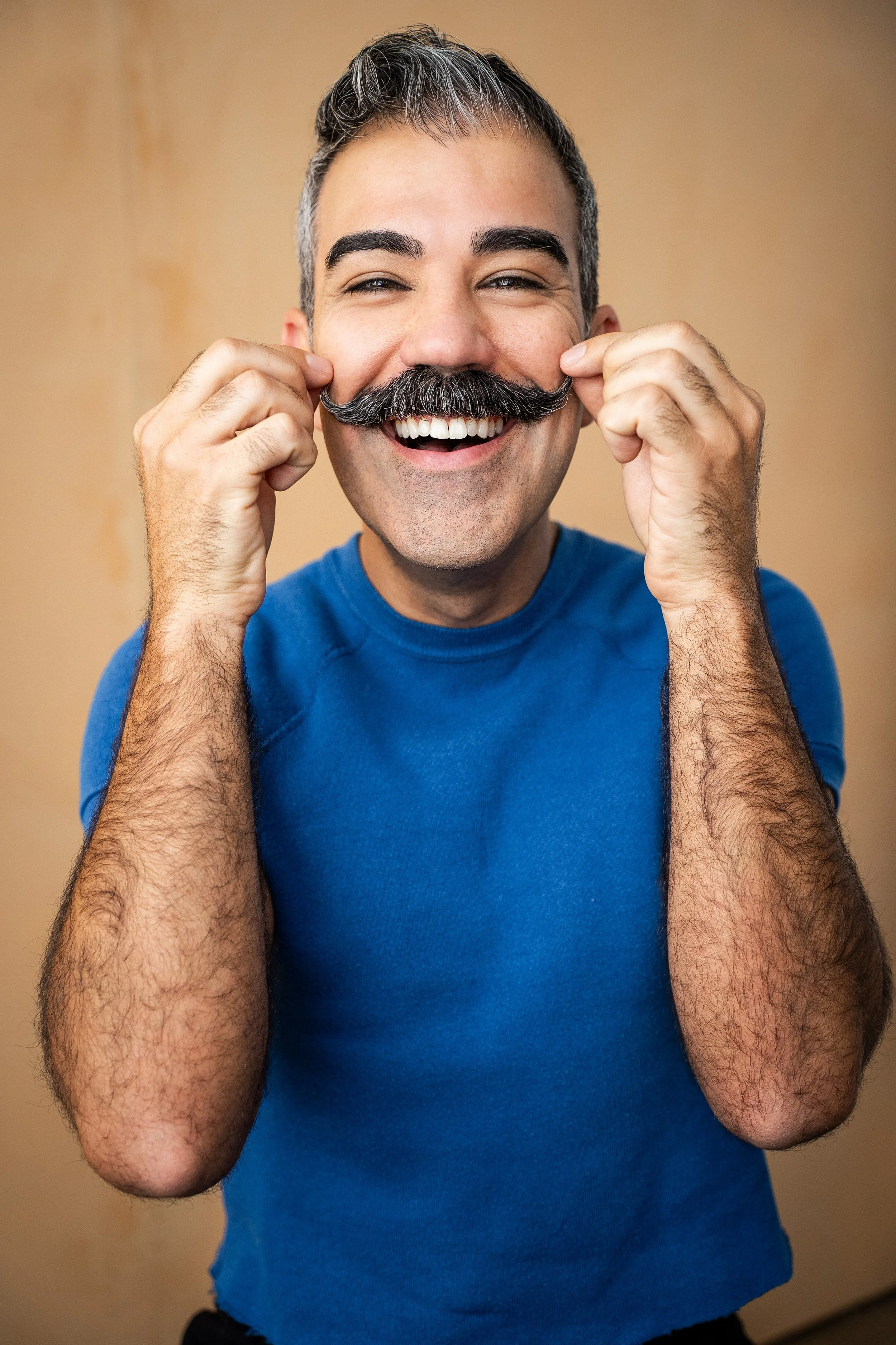 A smiling man with a thick mustache, gray hair, and a blue shirt, holding his moustache with both hands in front of a beige background.