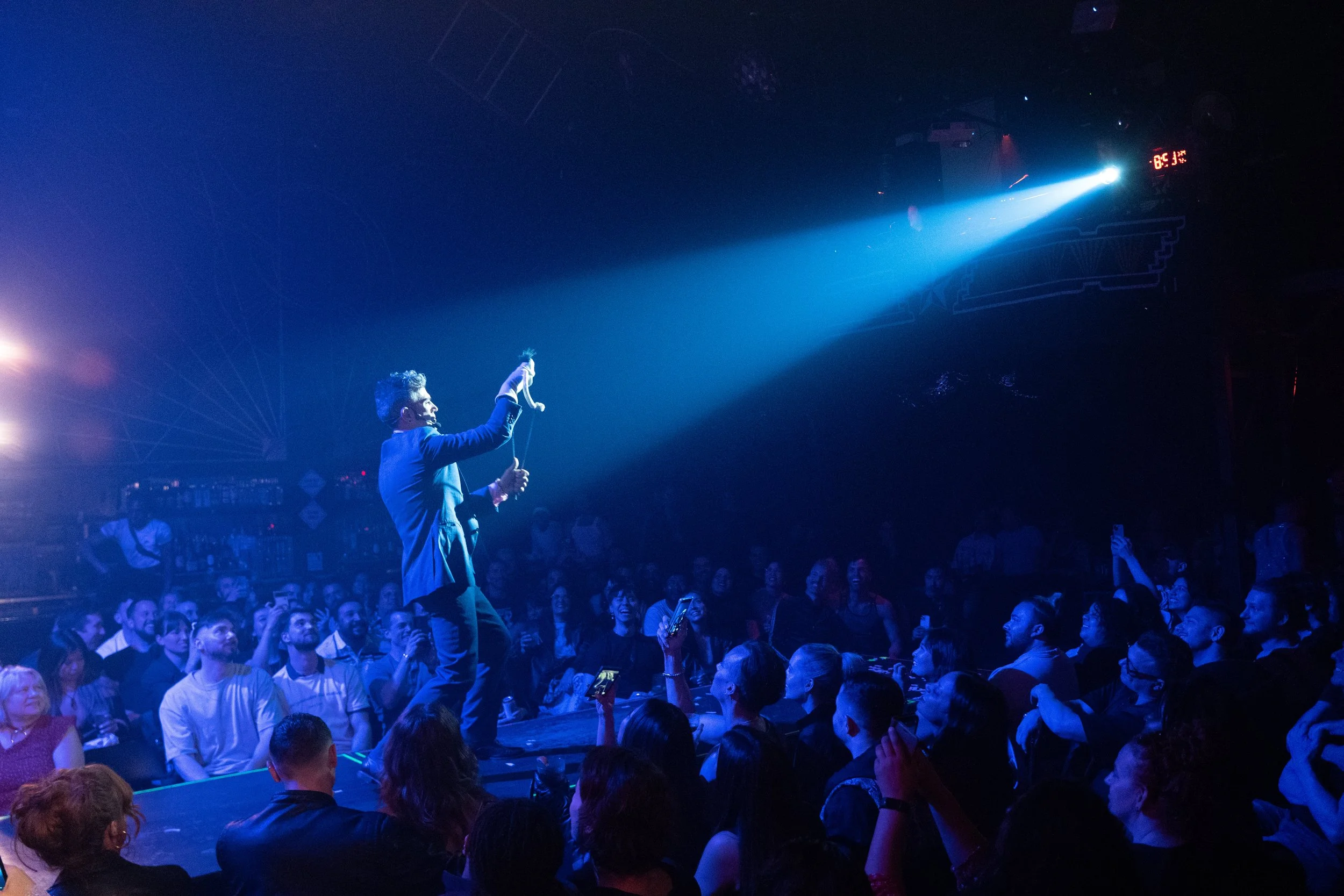A performer on stage with a microphone in hand, illuminated by a blue spotlight, entertaining a large audience in a dimly lit venue.