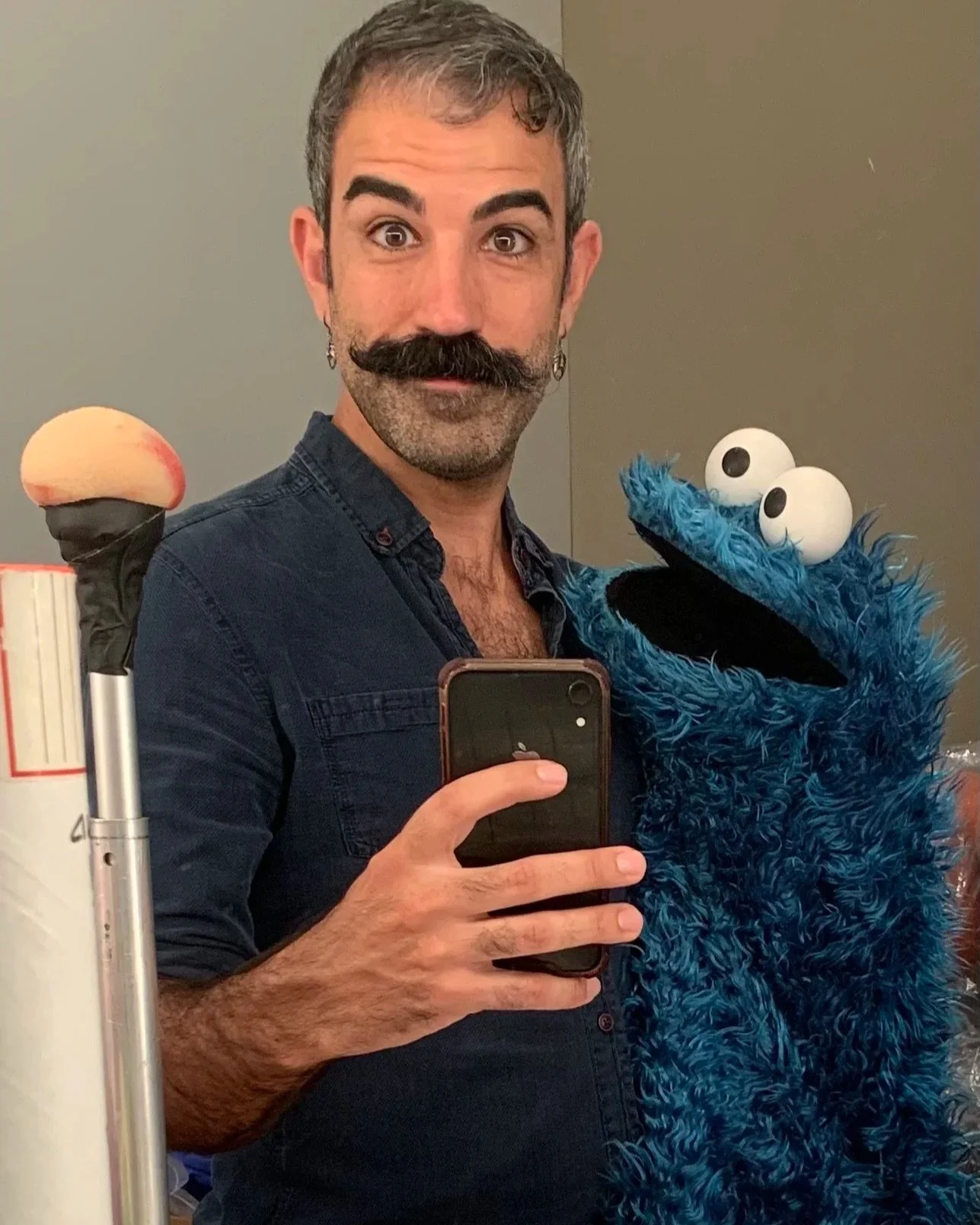 A man with a mustache and earrings taking a selfie with a blue furry puppet with big eyes, in a room with plain walls.