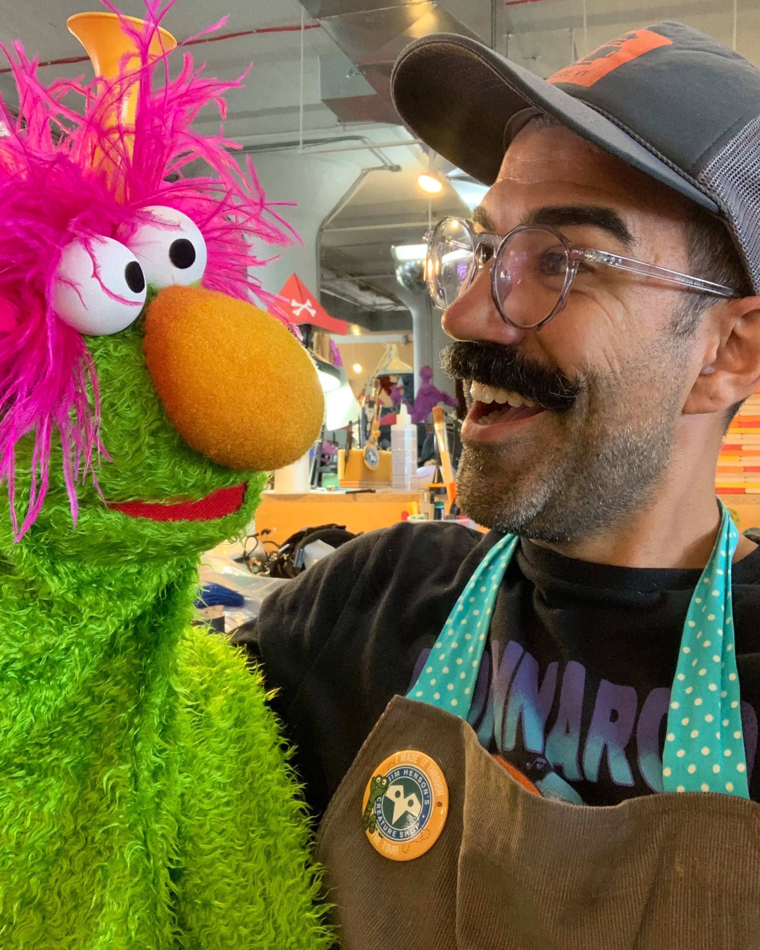 A man with glasses and a beard wearing a black shirt, a wide-brimmed hat, and an apron with a park badge, smiling and looking at a colorful puppet with vibrant pink hair, white eyes, an orange nose, and green fur.