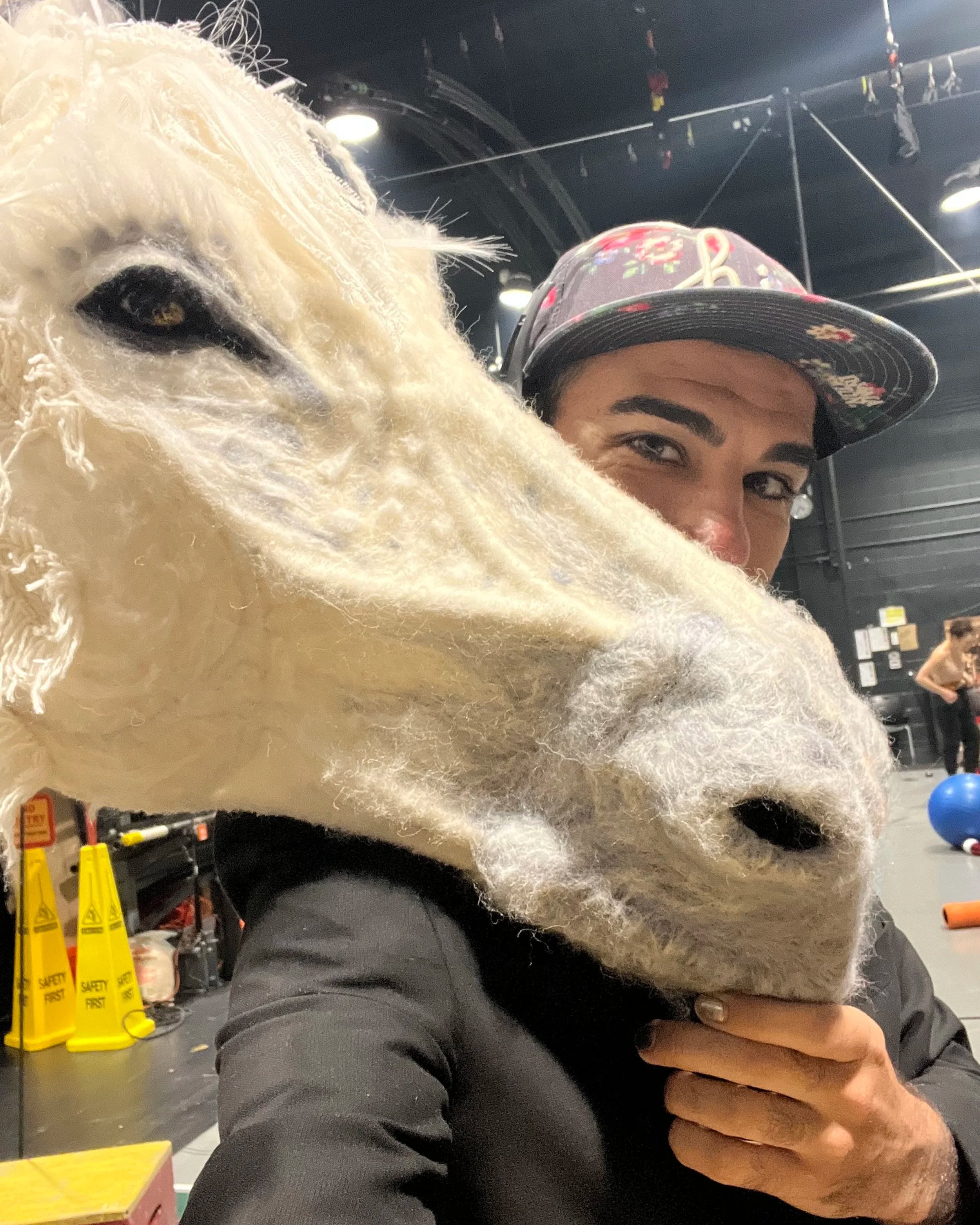 A woman with dark hair, wearing a floral cap, is hugging a large, fluffy white dog with a curly coat in an indoor setting, with caution signs and equipment visible in the background.