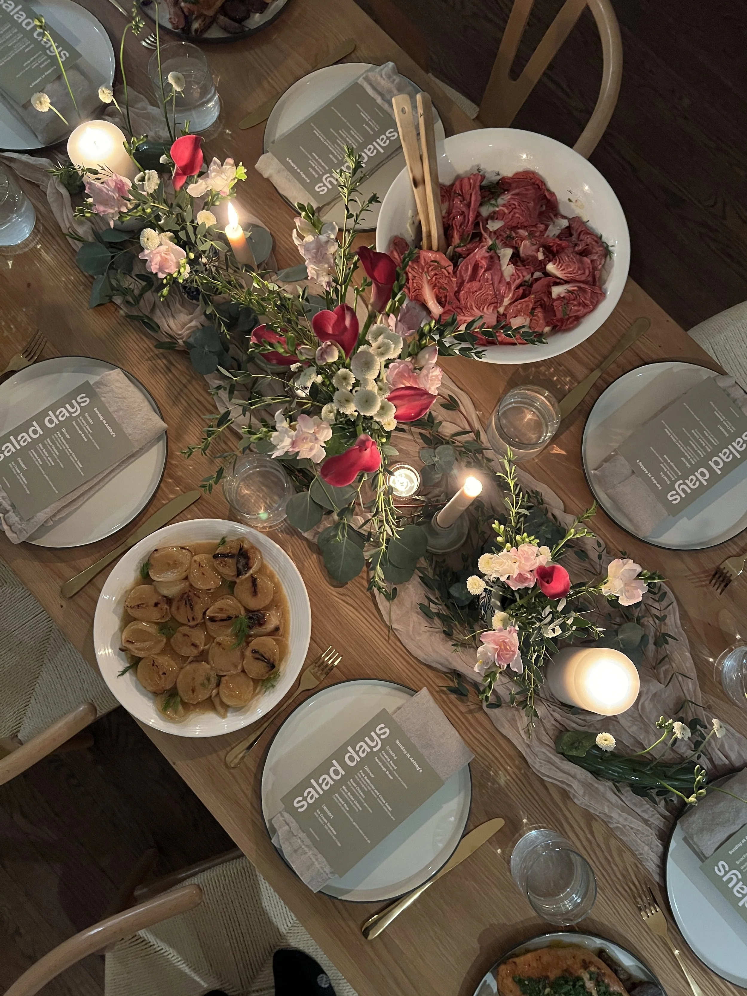 A dining table set with a floral centerpiece, lit candles, plates with salads, a bowl of sliced cured meat, and glasses of water, for a gathering or special occasion.