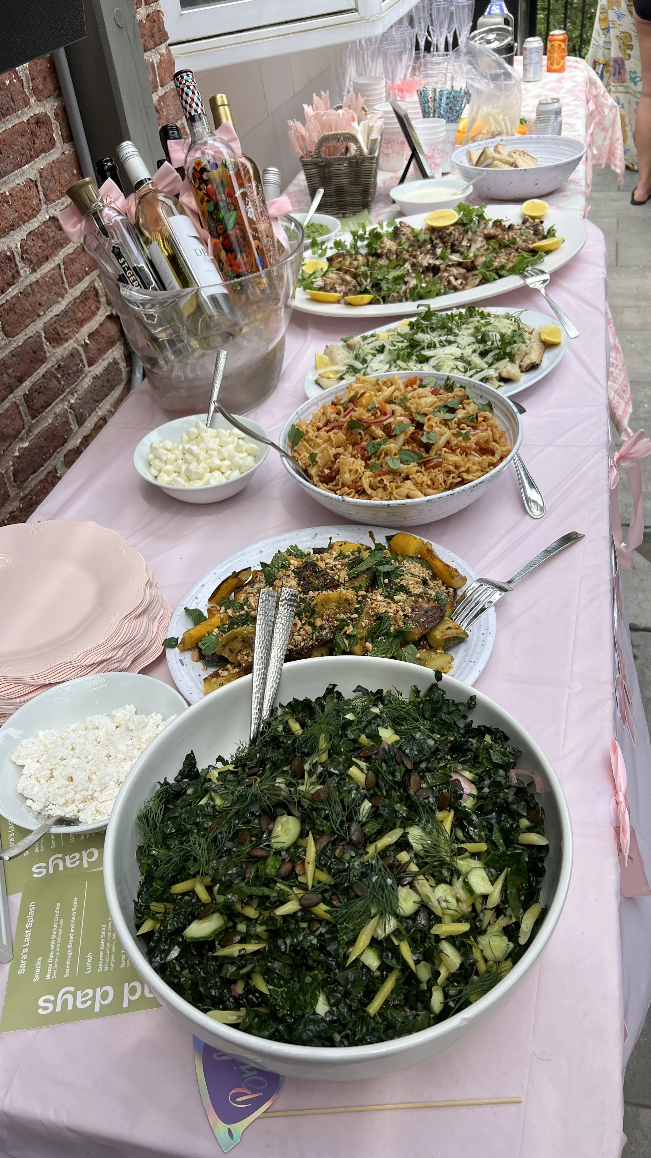 A table with various dishes including a green leafy salad, pasta salad, roasted vegetables, and grilled meat, with beverages and utensils for a gathering or party.