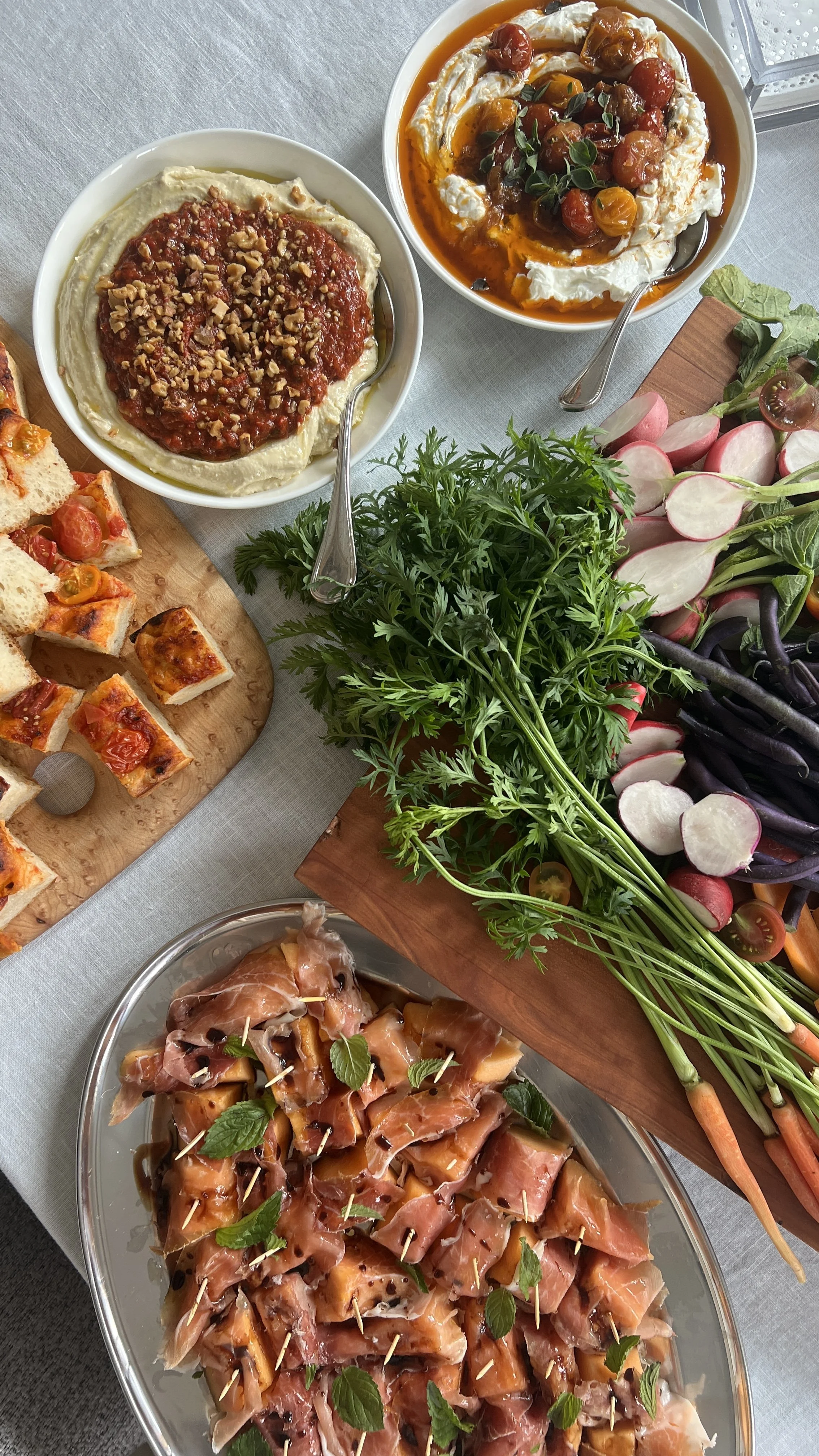 A variety of appetizers and fresh vegetables on a table, including a platter of prosciutto-wrapped melon bites garnished with basil, a wooden board of assorted mini pizzas, bowls of hummus with chopped nuts and cherry tomato topping, a bowl of cherry