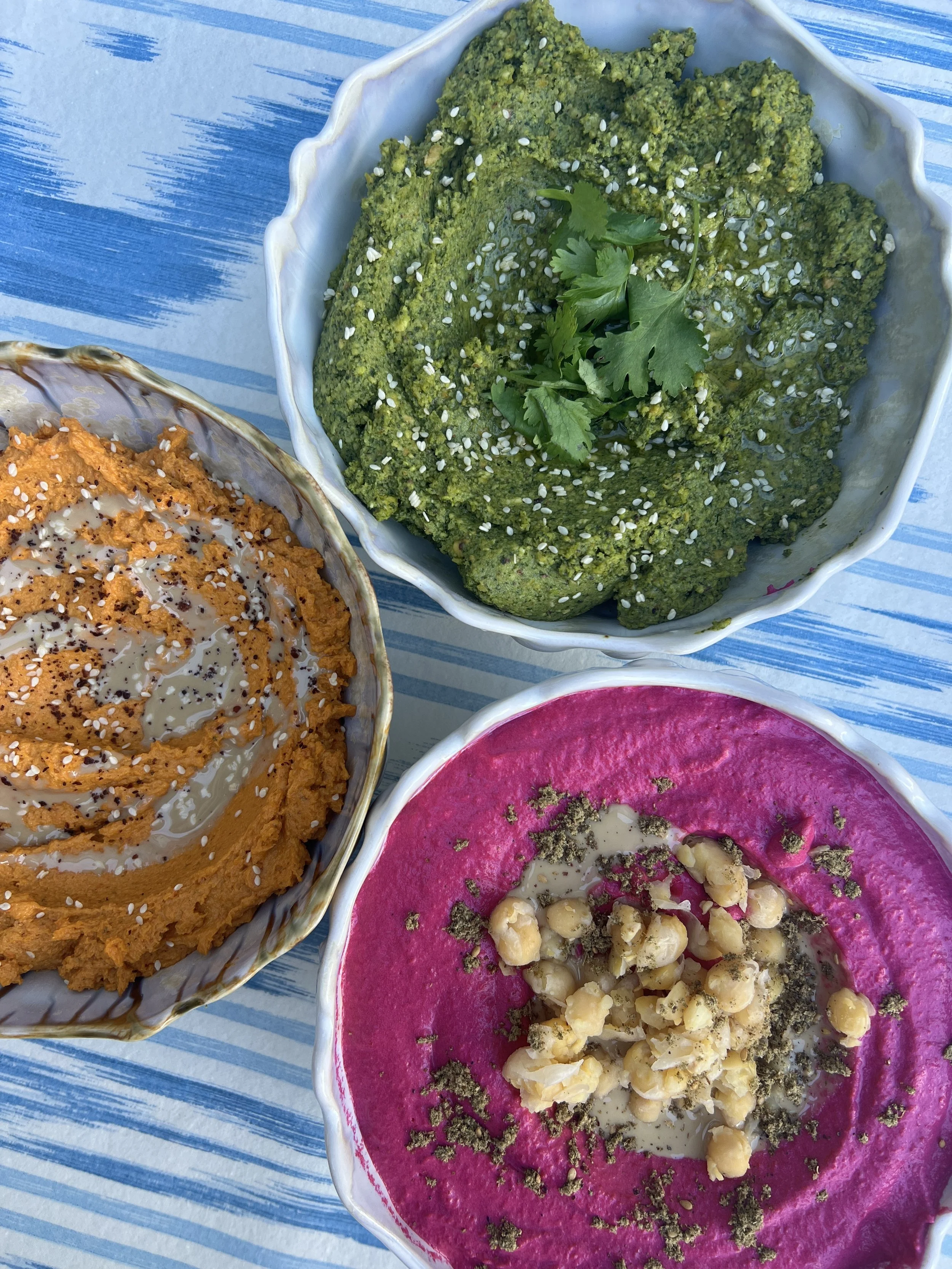 Three bowls of colorful hummus dips garnished with herbs, seeds, and spices on a blue striped tablecloth.
