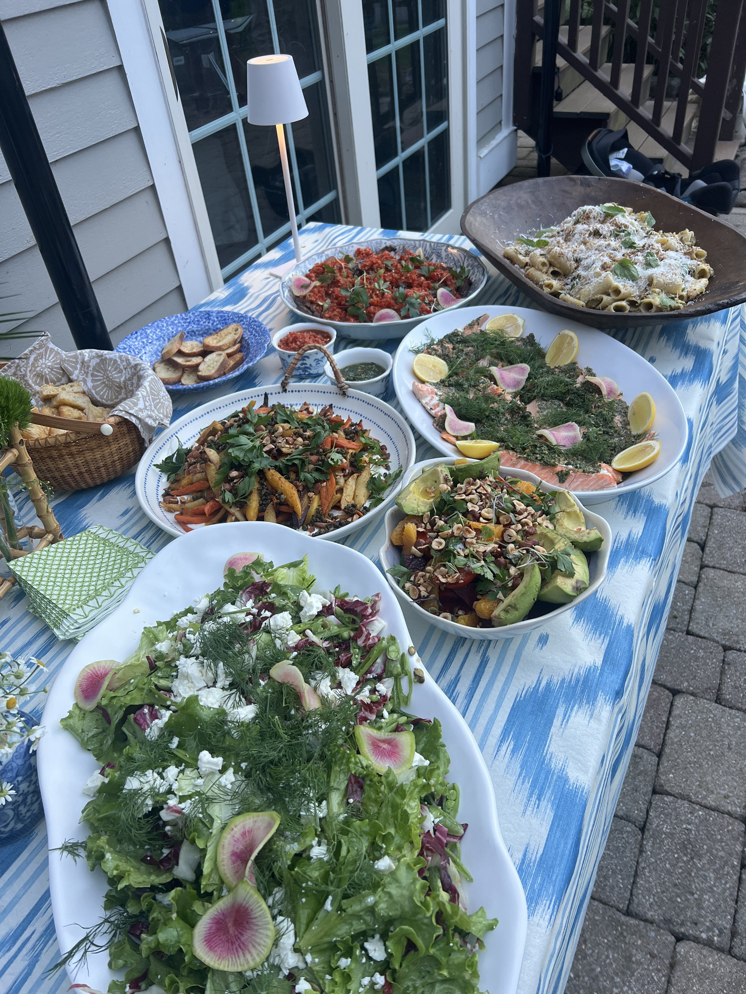 A table set with various dishes including a green salad with watermelon radish slices, a pasta dish with creamy sauce, a grilled salmon salad with lemon slices and herbs, a vegetable stir-fry, a tomato and herb salad, and a bowl of baked pasta, with 