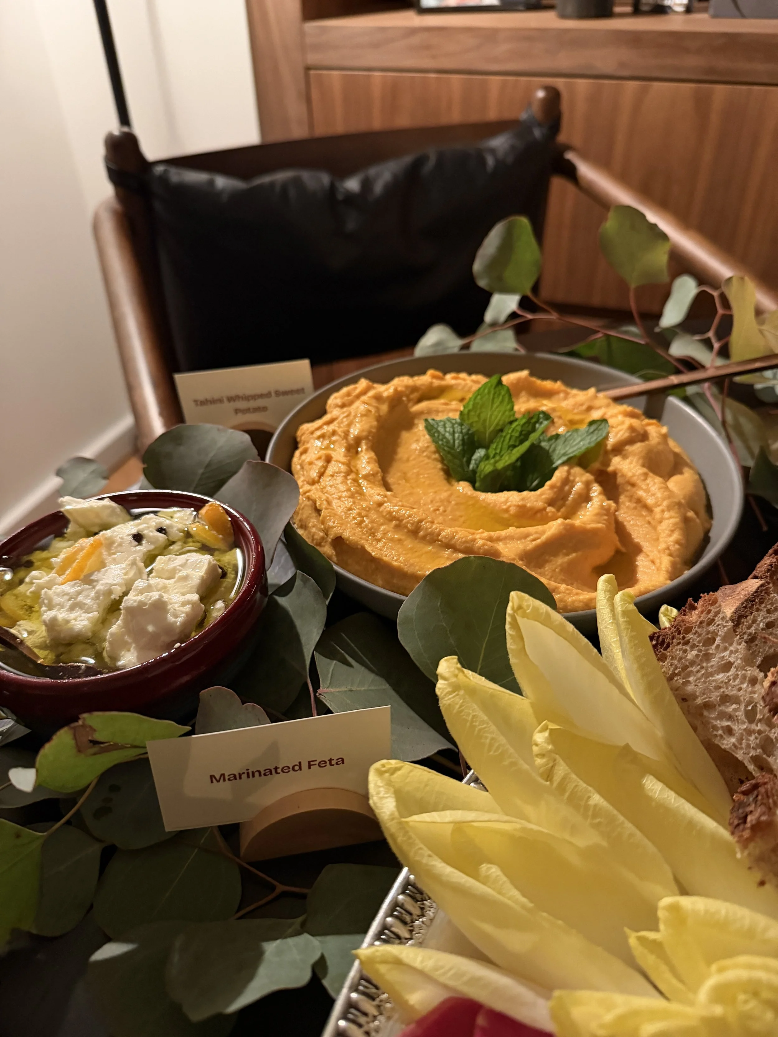 A spread of Middle Eastern dishes including hummus topped with mint, marinated feta cheese, and fresh yellow lemon wedges, with a background of a wooden chair and cabinet.