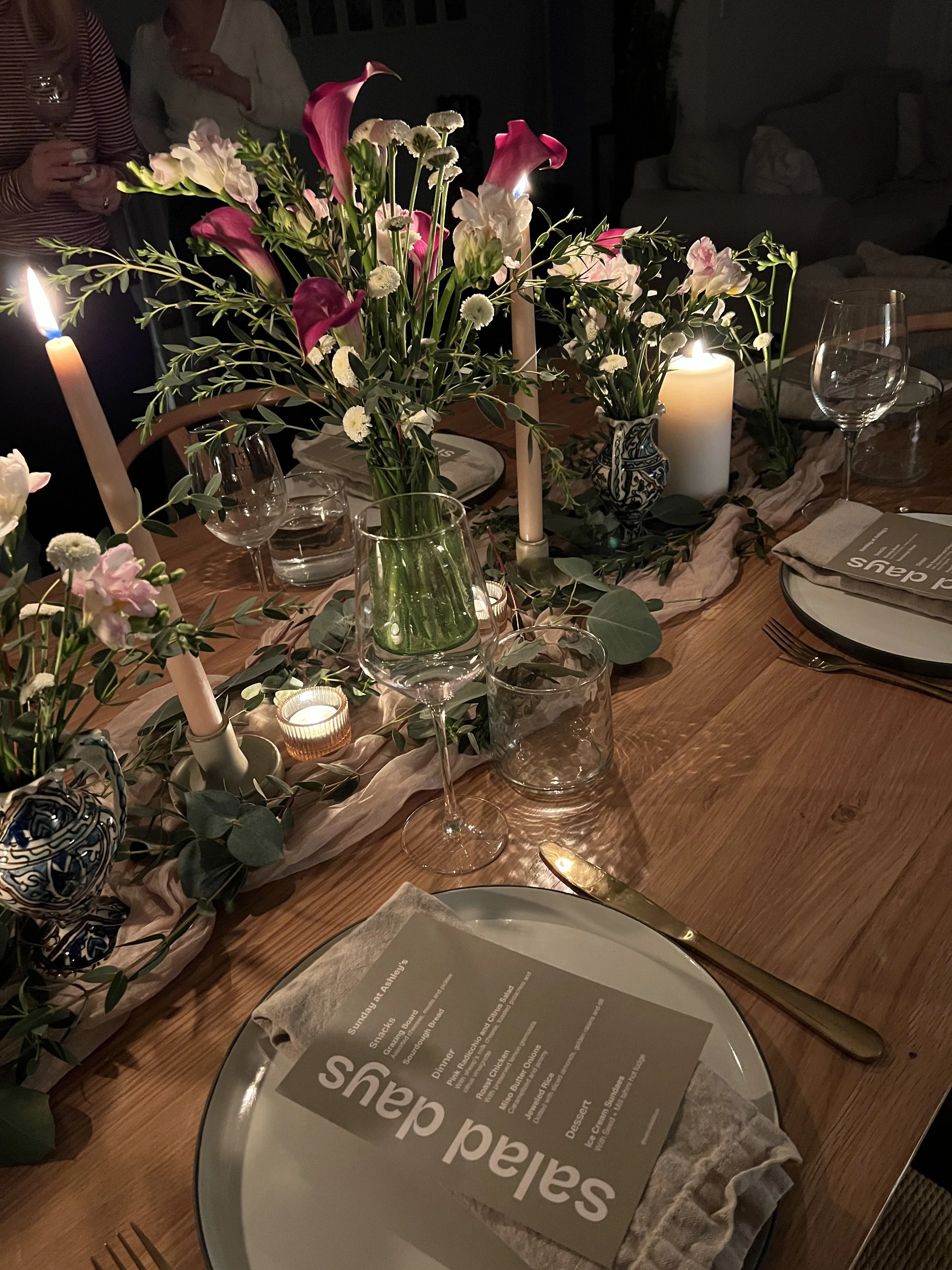 A dinner table decorated with pink and white flowers, candles, wine glasses, plates, and utensils, set for a special occasion.