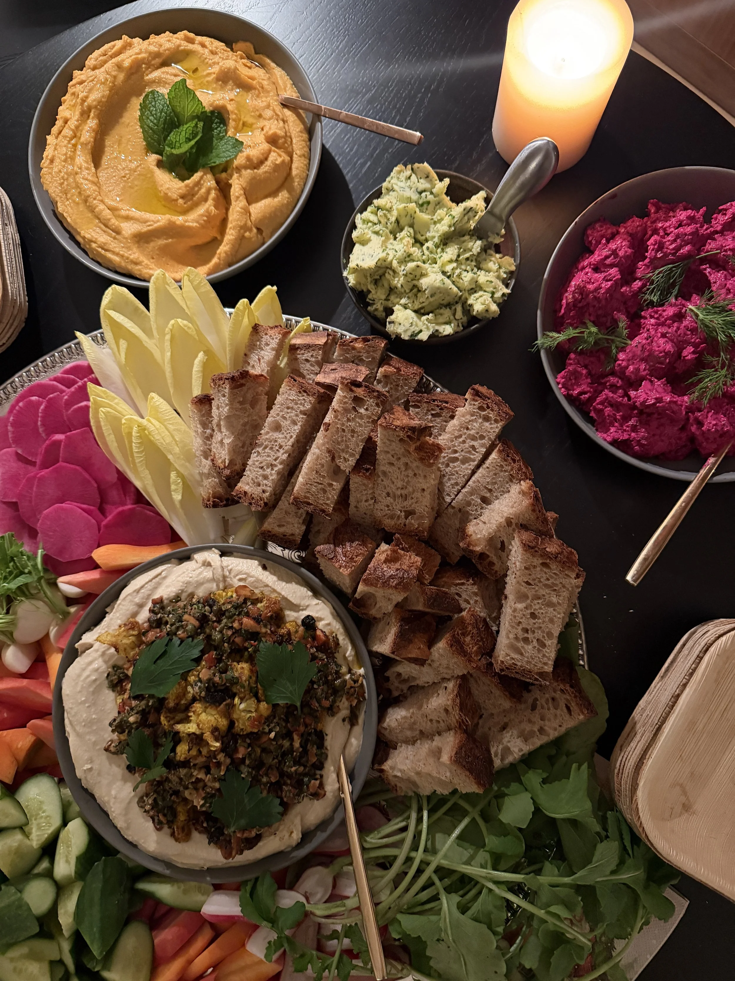 Assorted Middle Eastern dishes including hummus topped with herbs, guacamole, beet salad, pita bread, baba ganoush, vegetable sticks, and fresh greens, with a lit candle on a black table.