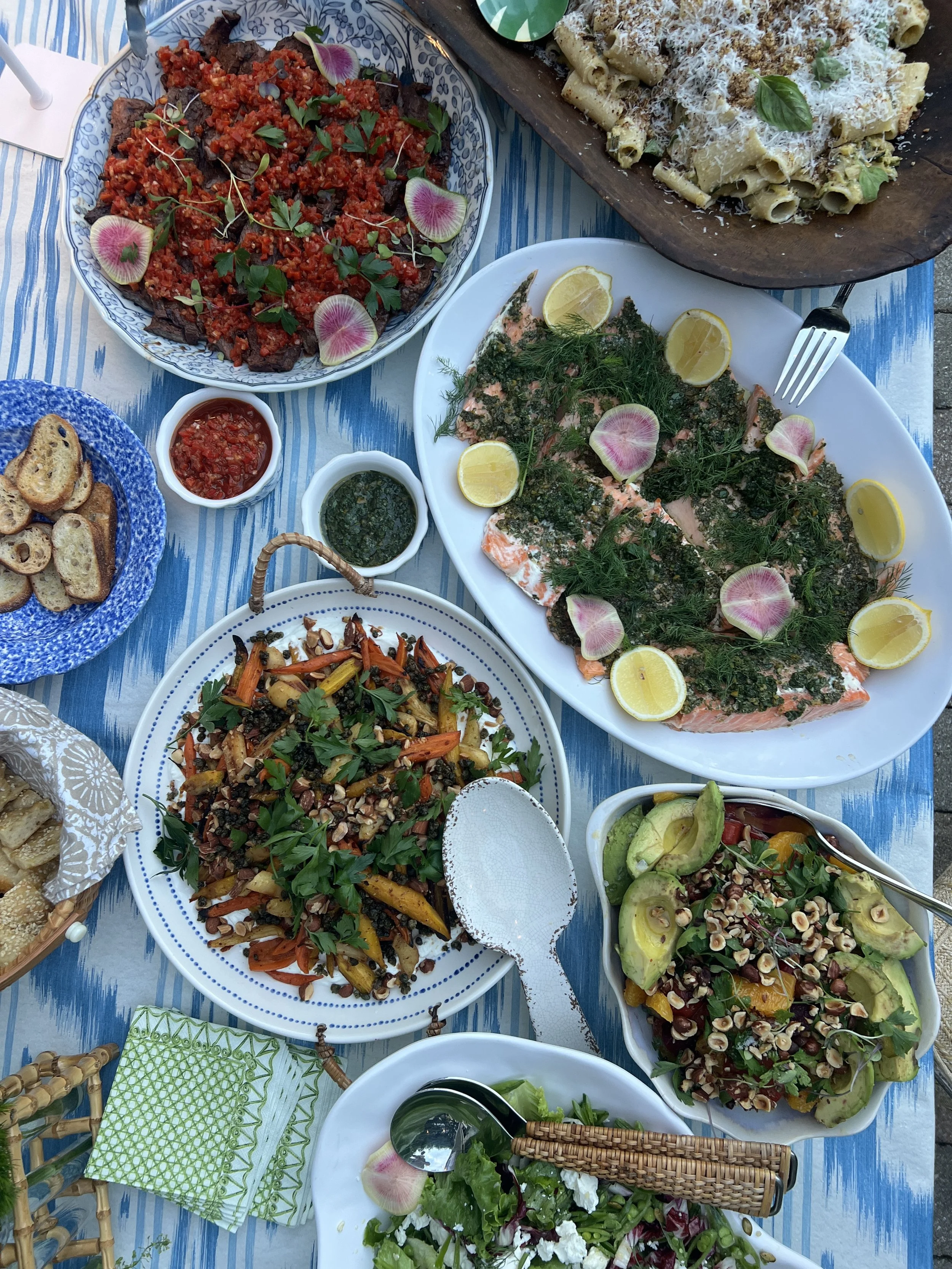 A variety of colorful dishes including a salad with greens, tomatoes, and grains, grilled salmon topped with herbs and lemon slices, pasta with cheese, roasted vegetables with herbs, avocado salad with nuts and fruit, and grilled bread, all arranged 