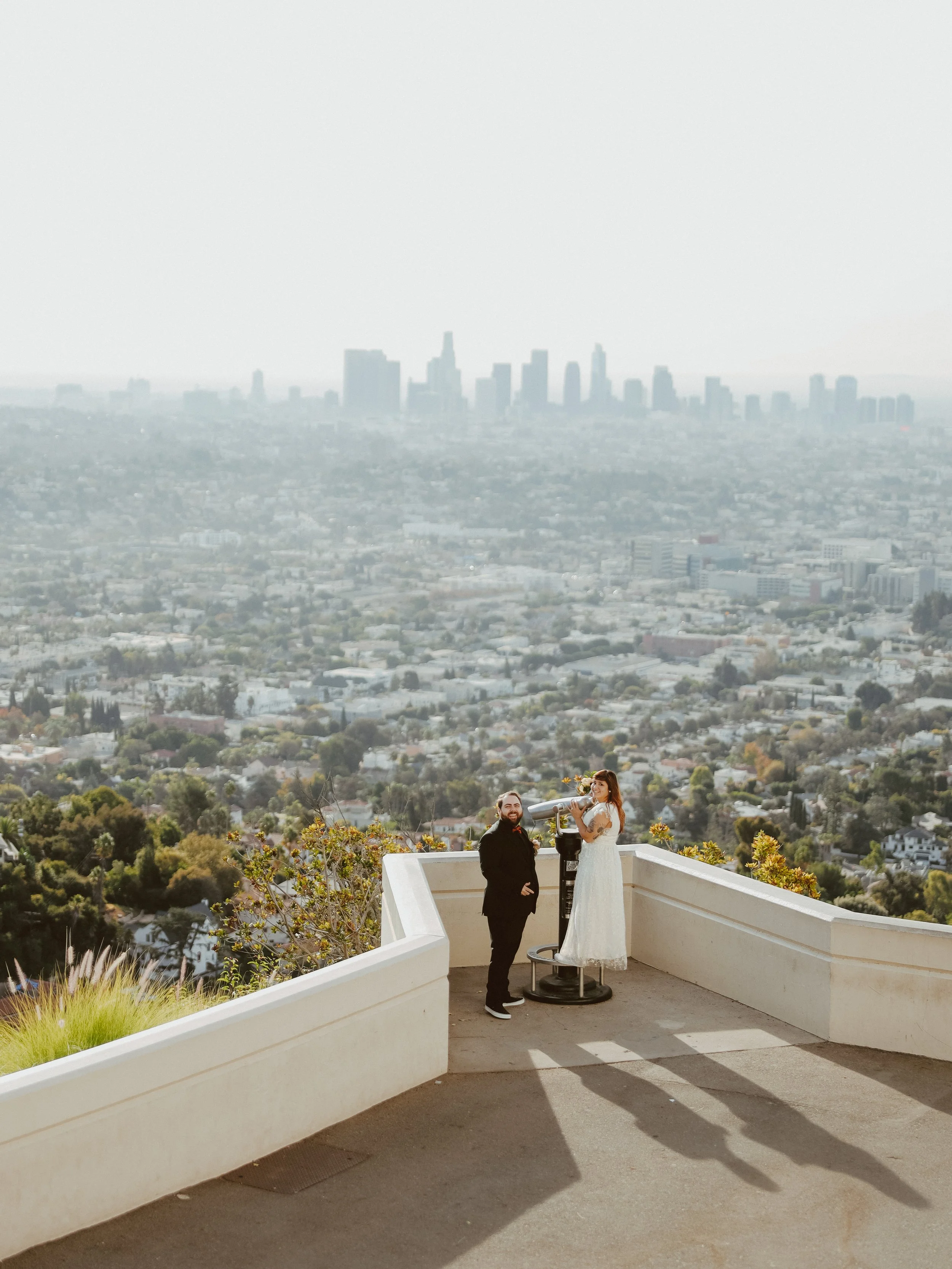 A couple standing at a scenic overlook with a large city skyline in the background, one person in a wedding dress and the other in a black suit, with a view of Los Angeles in the distance.