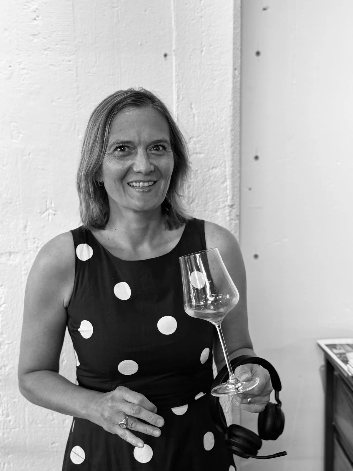 A woman smiling, holding a wine glass, wearing a polka dot dress, standing against a textured wall.