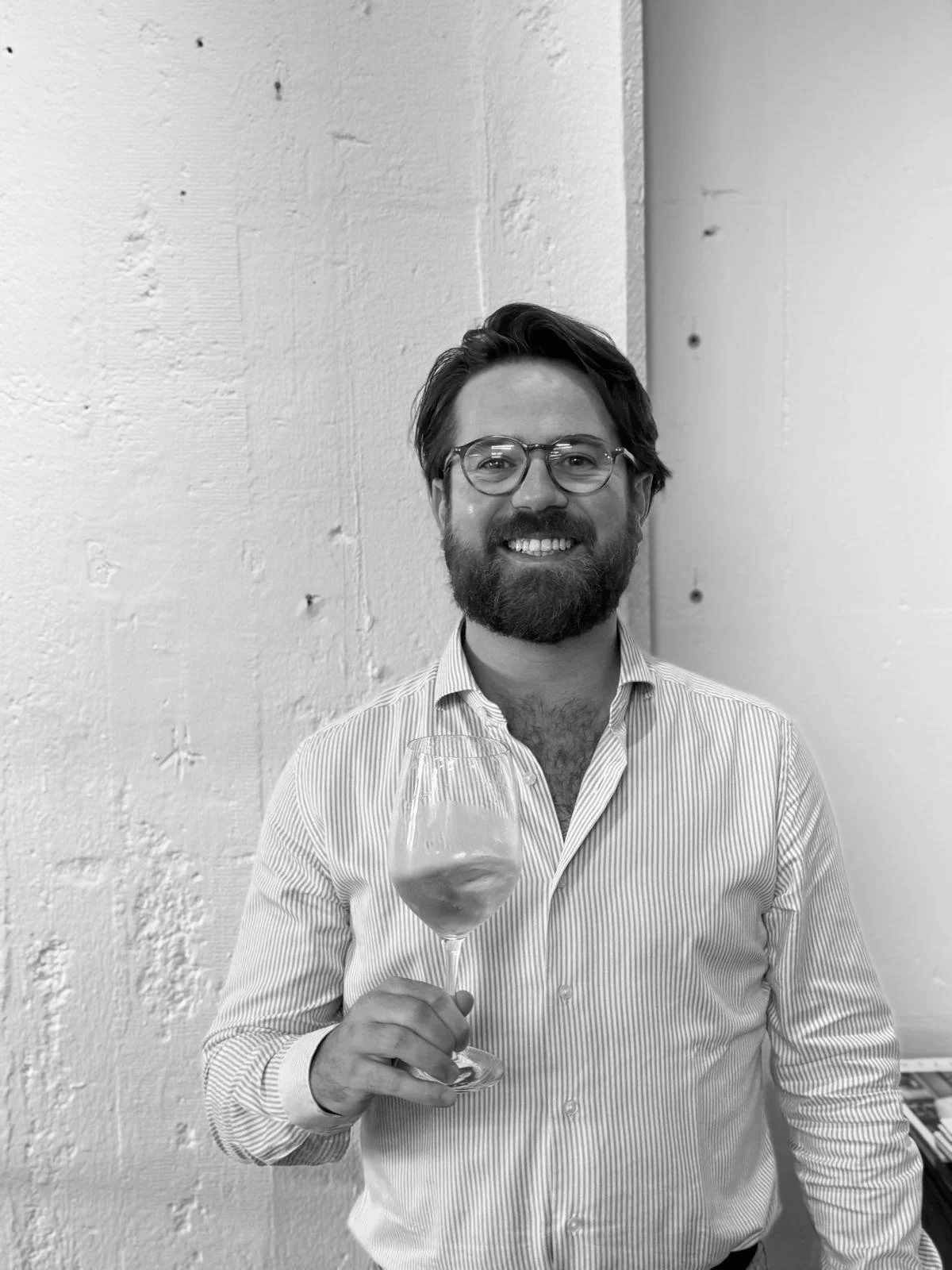 A man with glasses and a beard is smiling, holding a glass of wine, and wearing a striped shirt, standing against a textured wall.