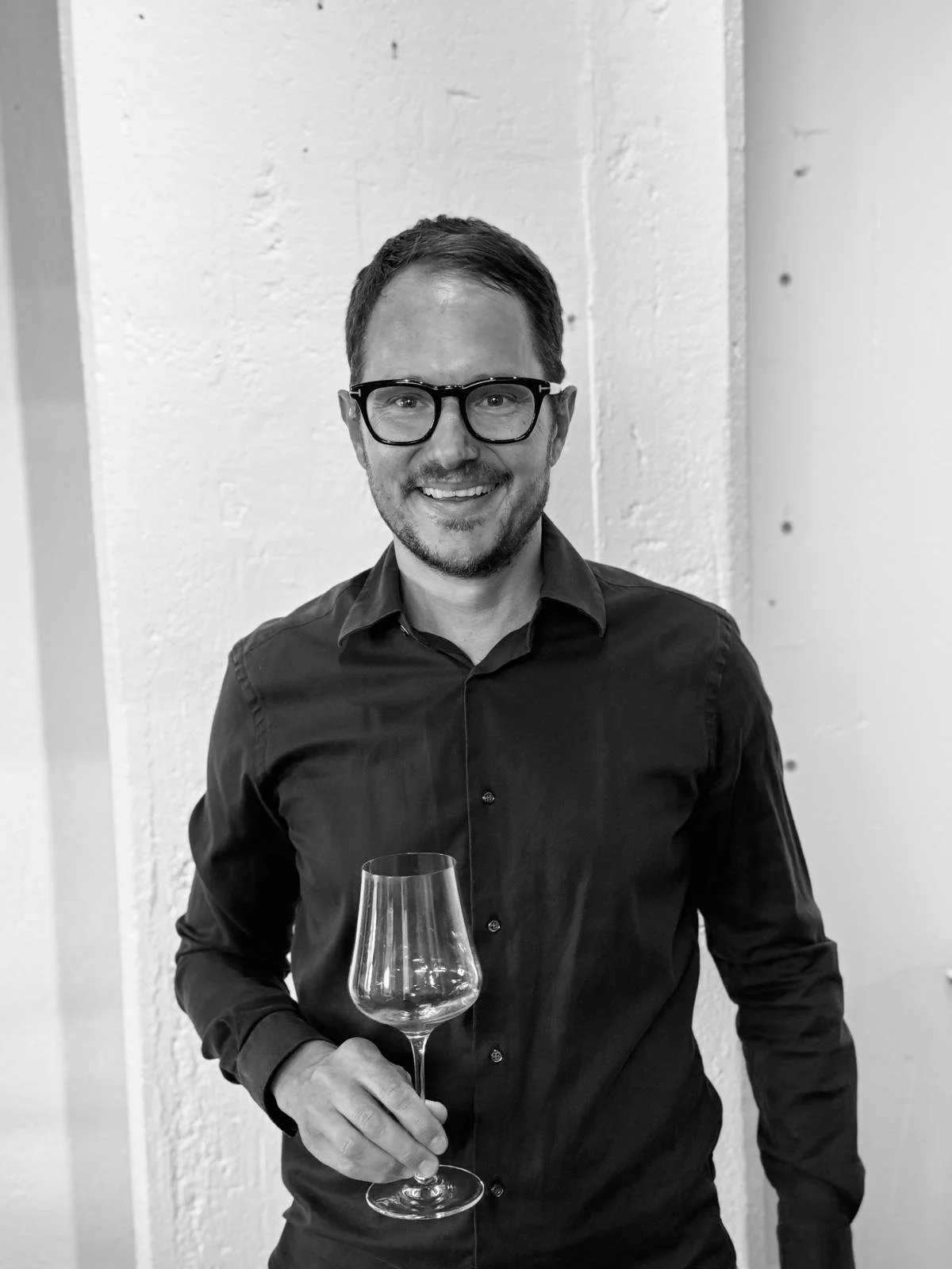 A man with glasses and a beard smiling while holding a wine glass, standing in front of a white wall.