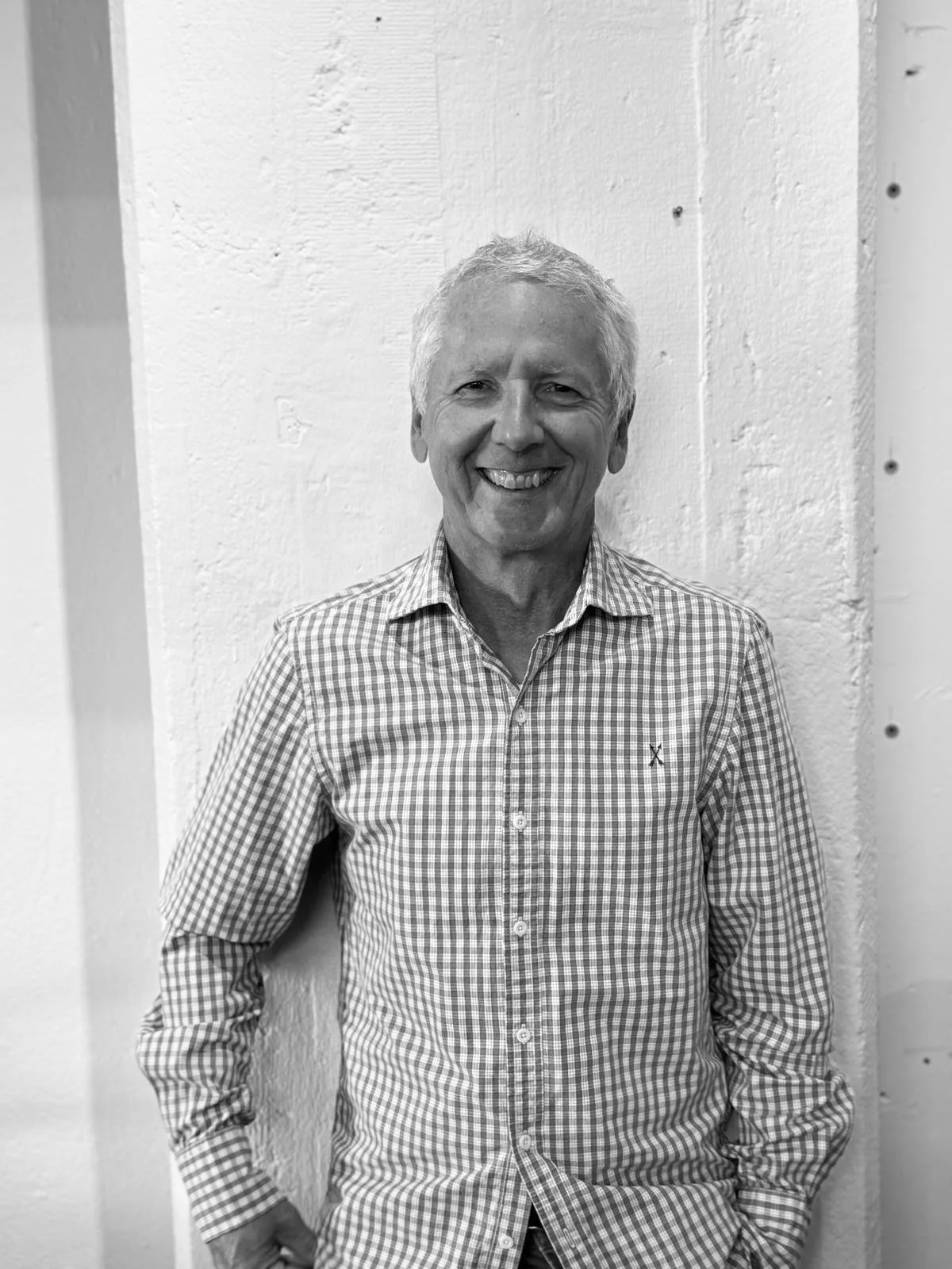 A smiling older man with gray hair wearing a checkered shirt standing against a textured white wall.