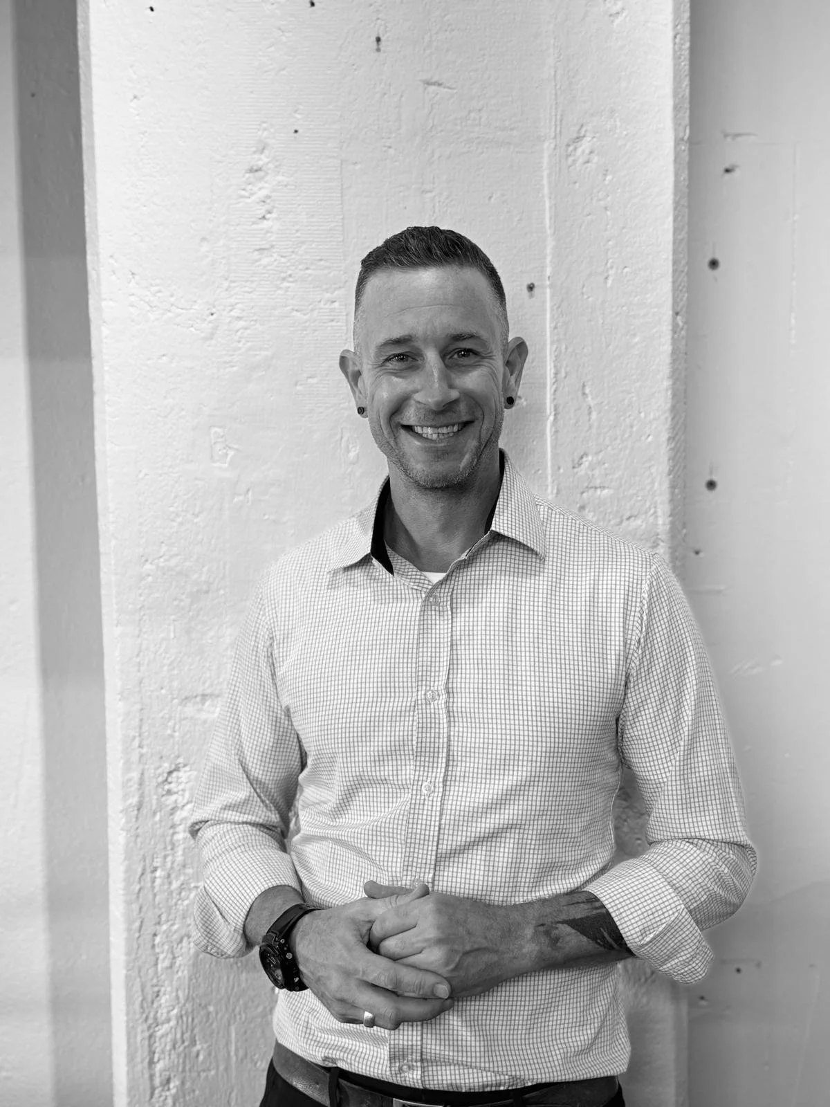 A smiling man with short hair, wearing a checkered shirt, standing against a white wall.
