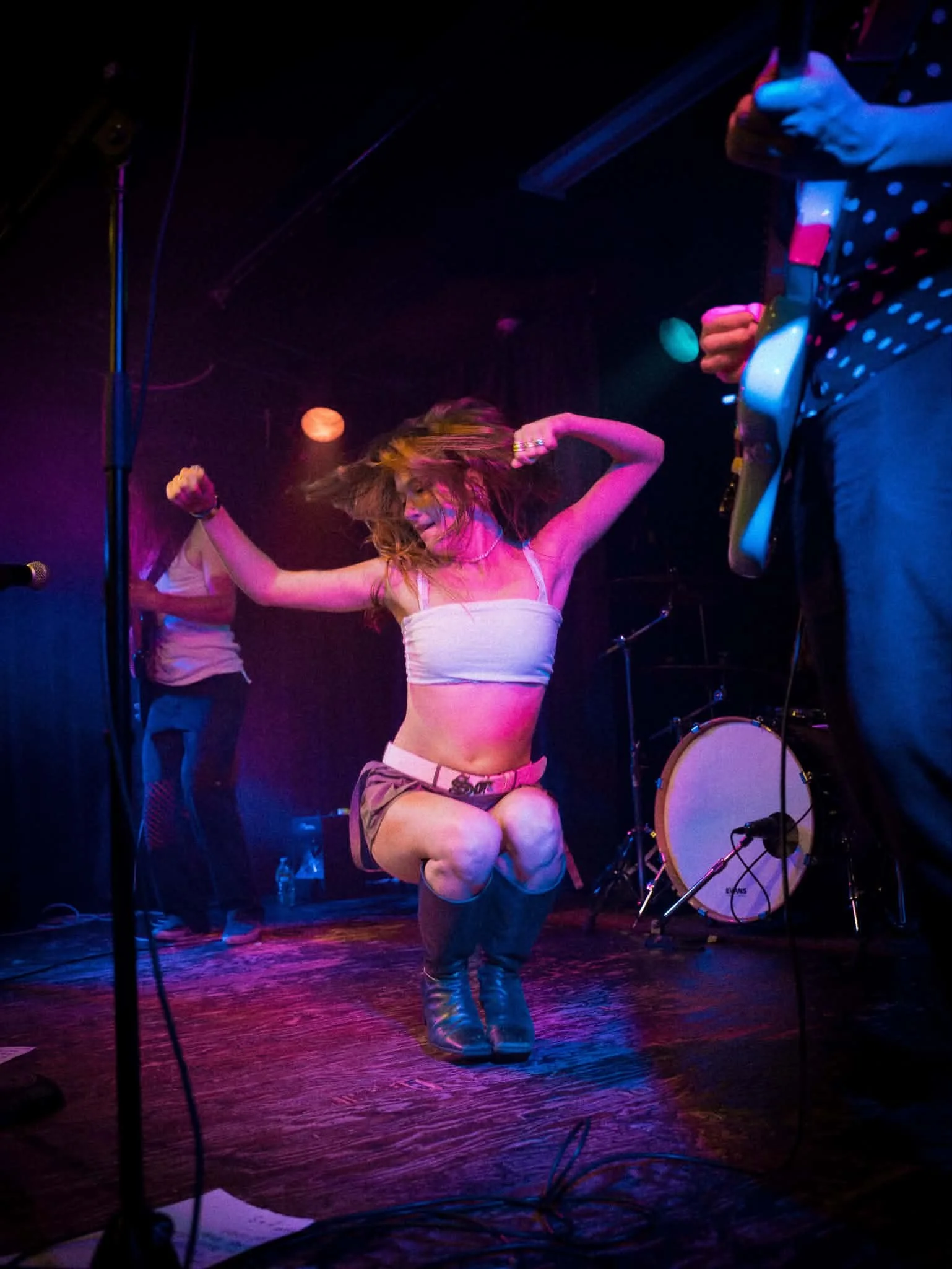 A woman dancing on stage in a white top and shorts, with colorful stage lighting, next to a musician playing guitar, with drums in the background.