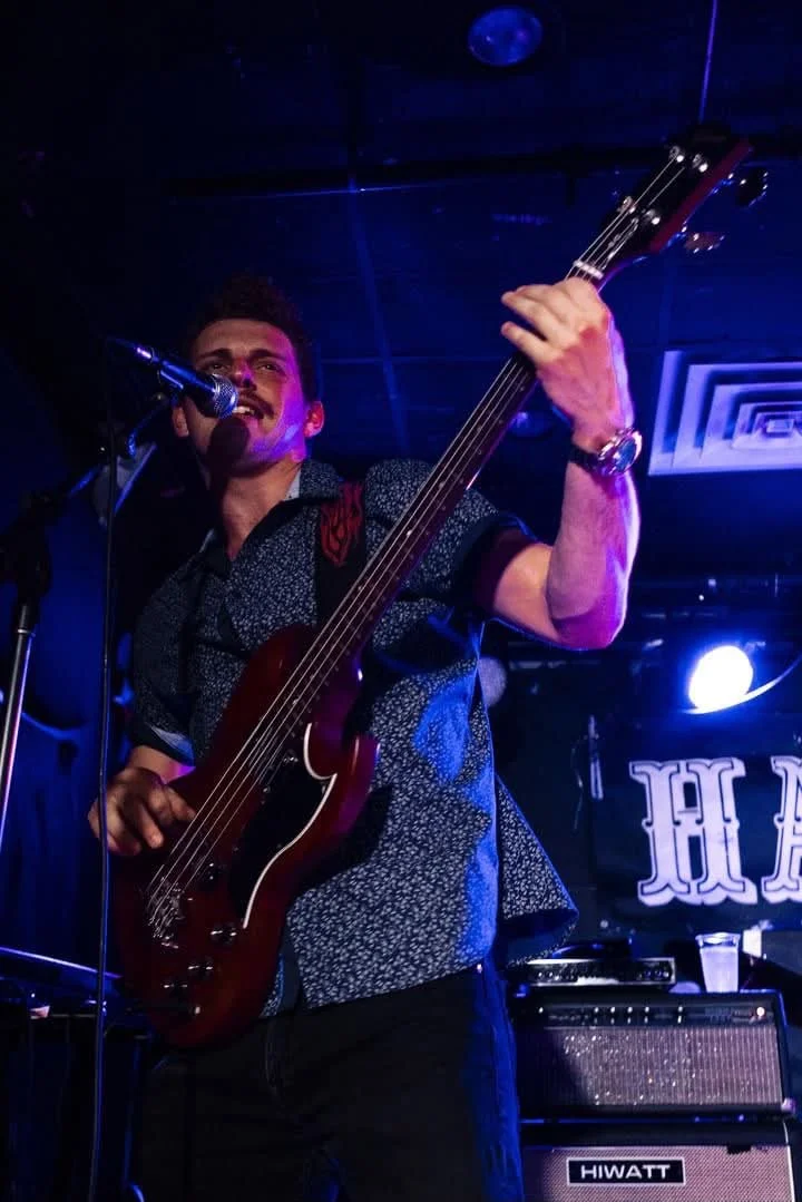Male musician playing an electric bass guitar and singing into a microphone on stage with blue and purple lighting.