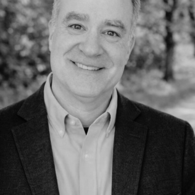 Black and white portrait of a smiling man in a suit jacket and collared shirt, outdoor background with trees.