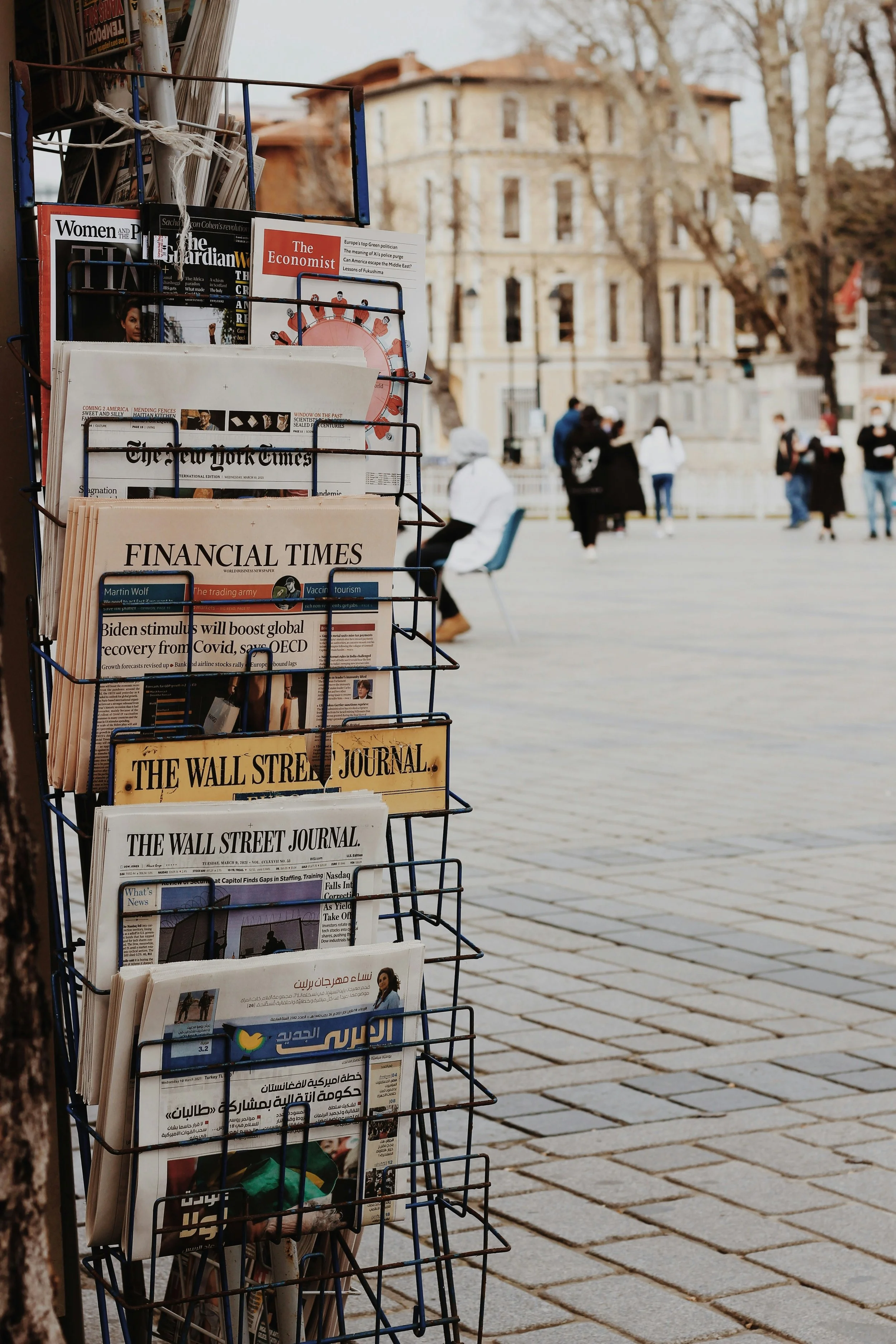 Free newspaper stand on a street with various newspapers and magazines including The Wall Street Journal, Financial Times, The New York Times, The Economist, and others in front of a historic building, with people walking and standing in the background.