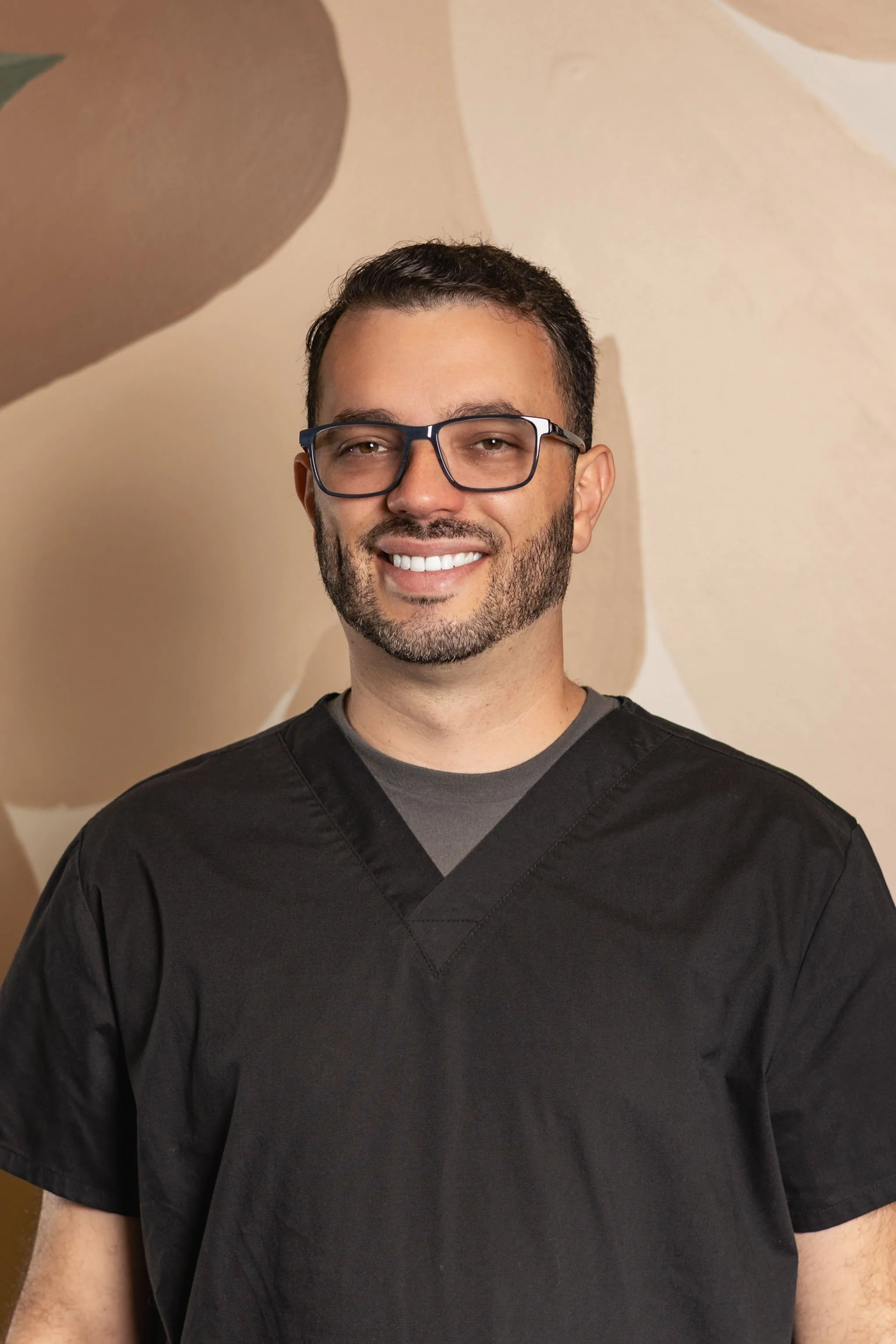 A smiling man with short dark hair, glasses, and a beard, wearing black scrubs, standing in front of a beige abstract wall.