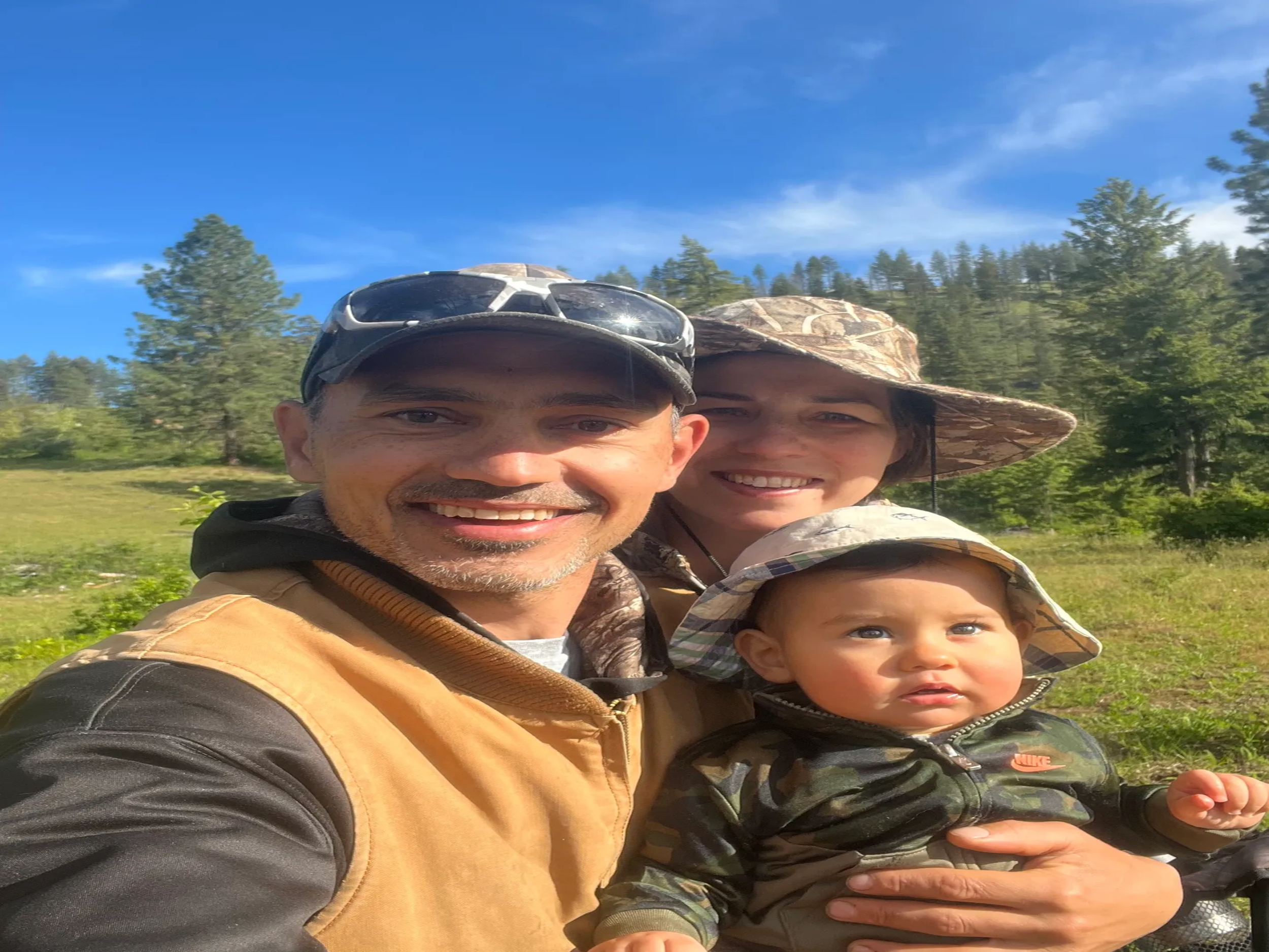 A family selfie outdoors with a man, woman, and baby wearing hats, surrounded by greenery and trees under a blue sky.