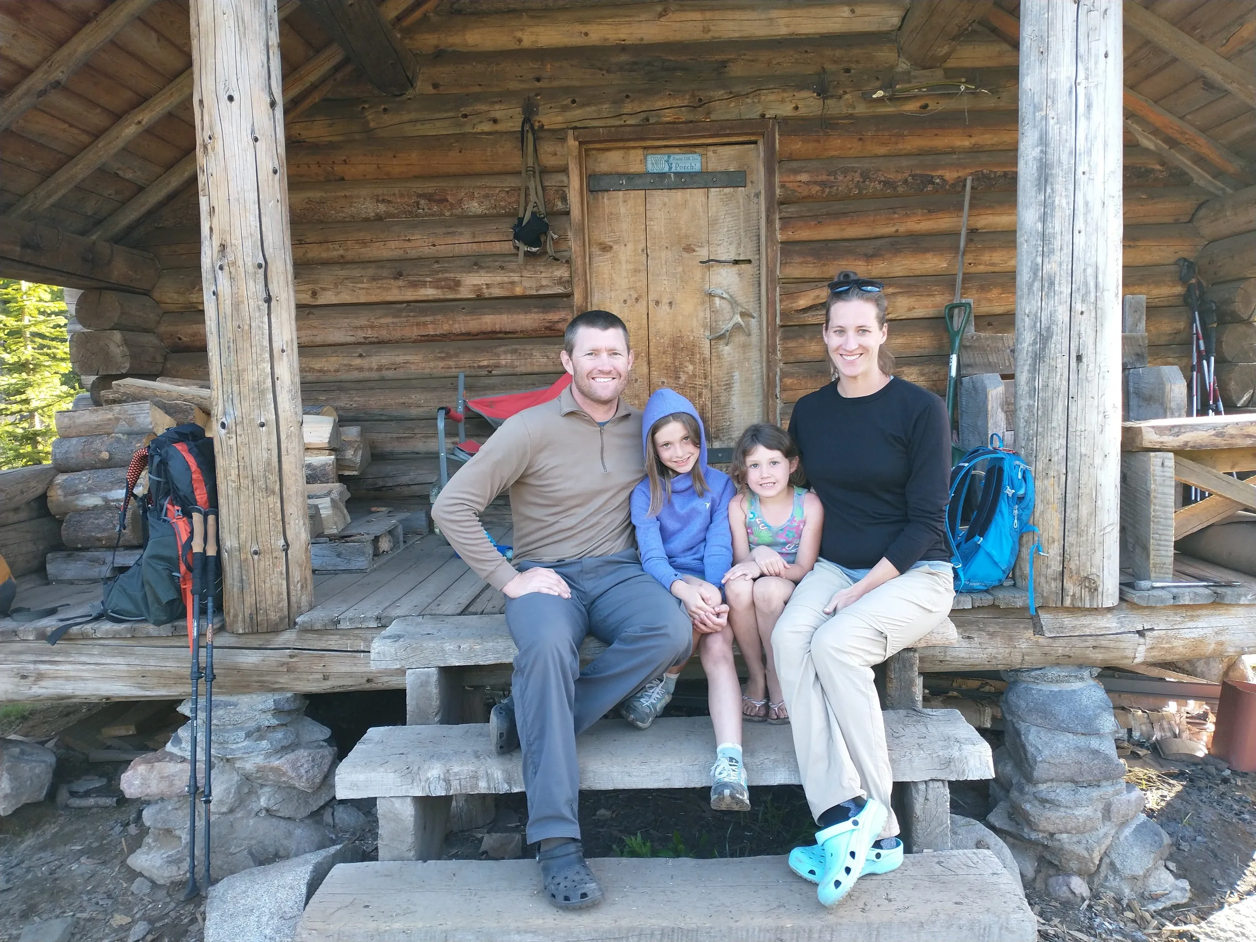A family sitting on the steps of a rustic log cabin. The group consists of an adult man, two children, and an adult woman. Hiking gear and trekking poles are visible on either side of the porch. The cabin is made of rough-hewn logs with a wooden door in the background.