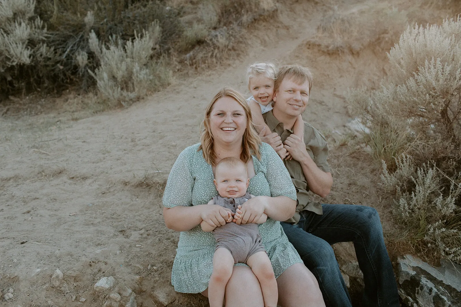 Family of four sitting outdoors with smiling parents, a baby in mother's lap, and a child on father's shoulders.