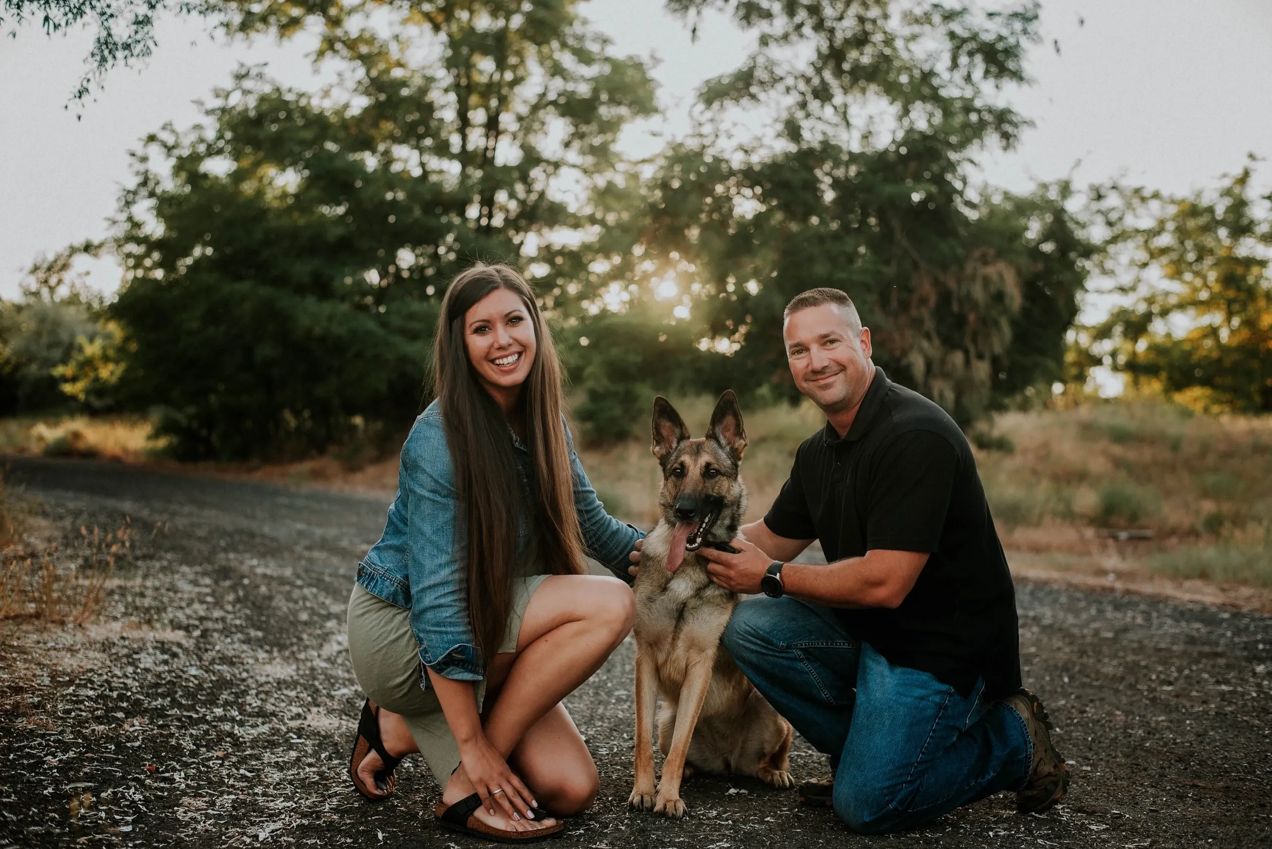 A man and woman kneeling on a path with a German Shepherd dog, surrounded by trees.