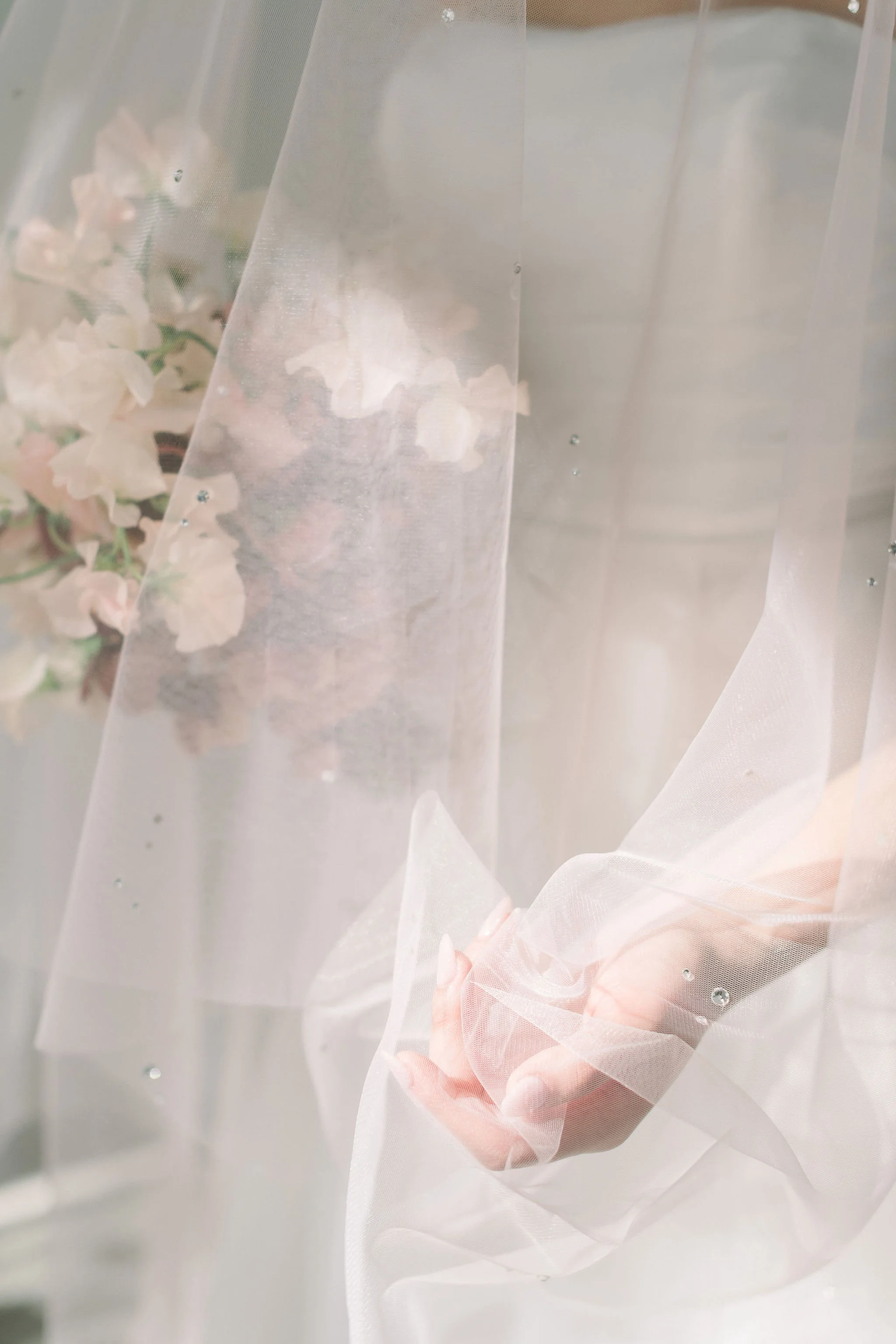 Close-up of a sheer Tessa Kim veil dotted with pearls, softly wrapping the bride’s hand and bouquet of blush sweet peas by Roots of Joy.
